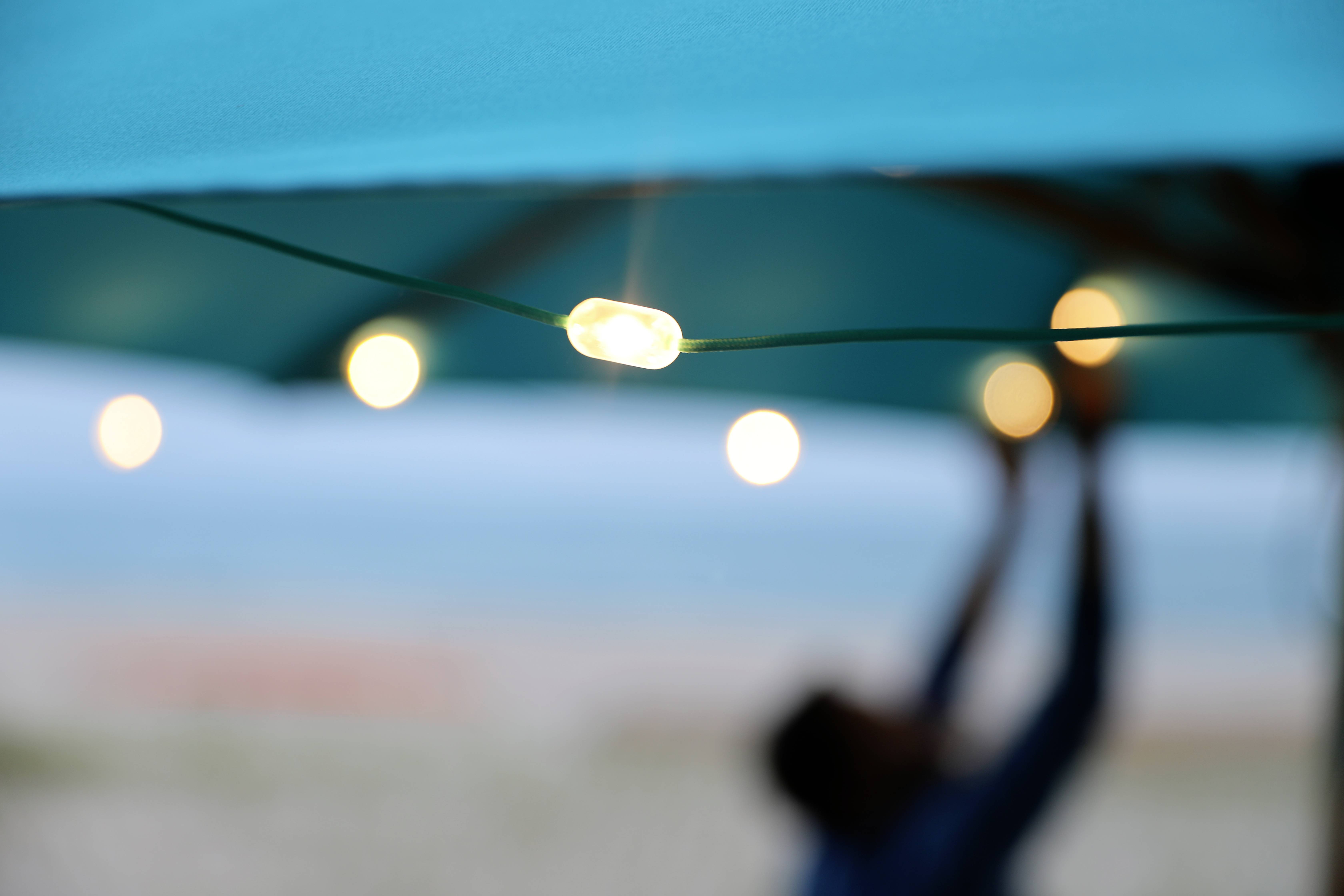 Onder een blauwe parasol hangt een rij van felle slingers; op de achtergrond een onscherp strand en zee.