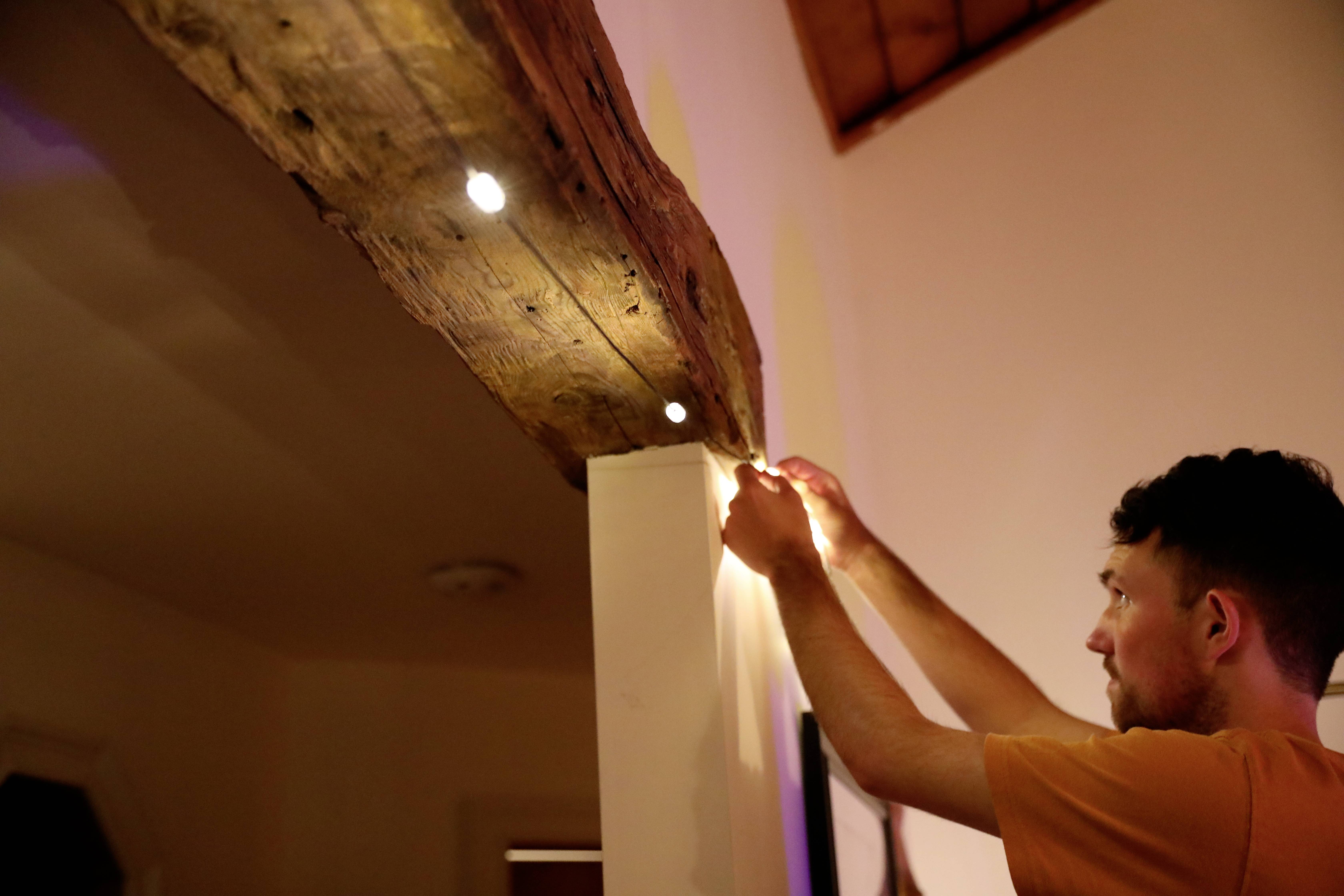 Een man installeert kleine LED-lampjes aan een houten balk in een ruimte met een houten plafond.