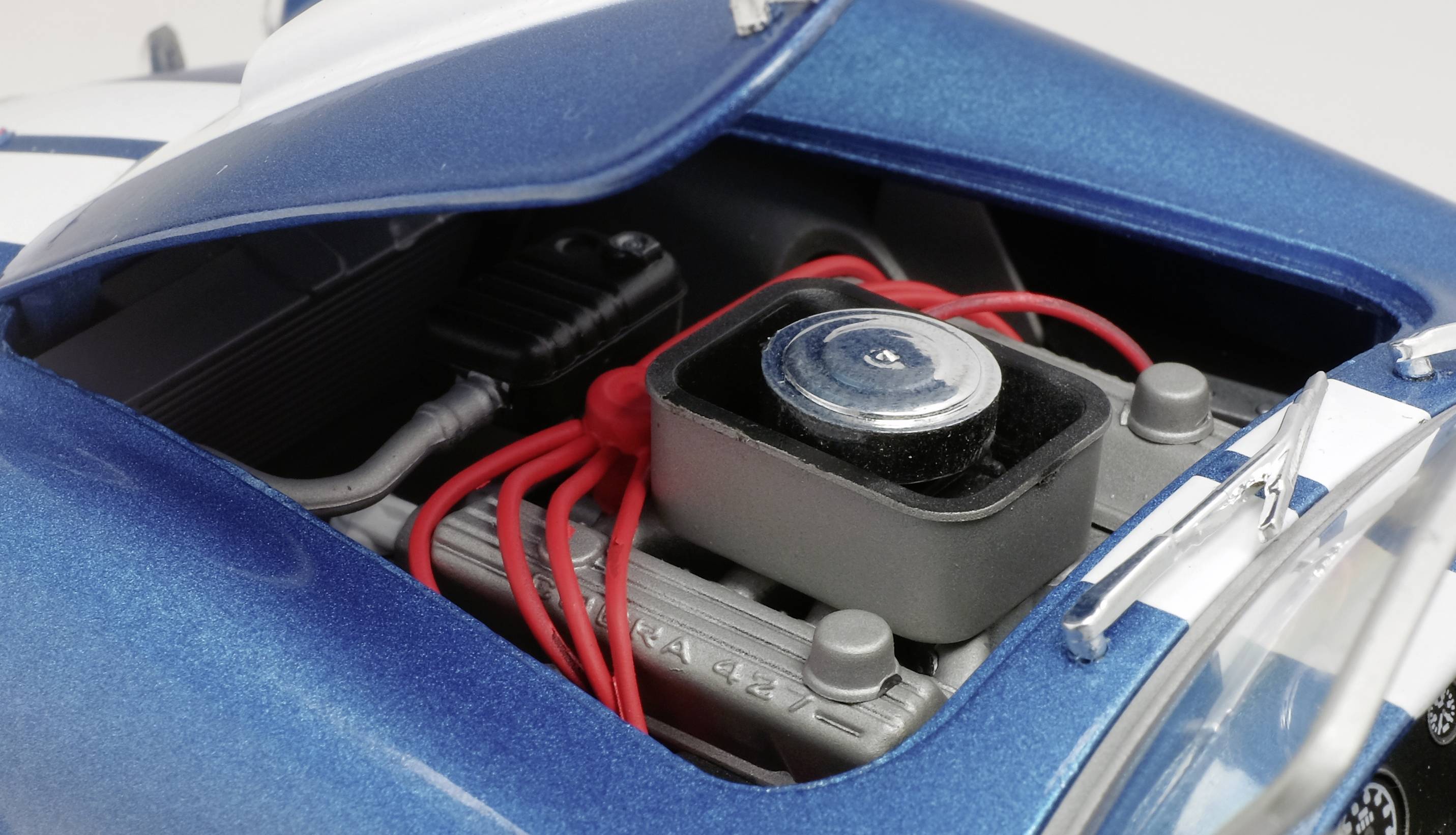 Close-up van een blauw modelautootje met een geopende motorkap, waarbij de gedetailleerde motor en rode ontstekingskabels zichtbaar zijn.