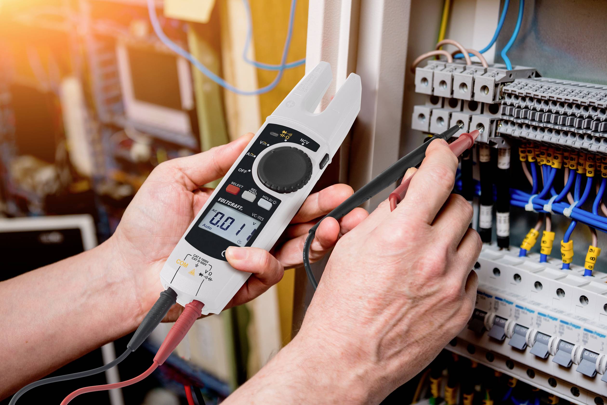 Een persoon meet spanning in een schakelkast met een digitaal multimeter. Kabels en zekeringen zijn zichtbaar op de achtergrond.