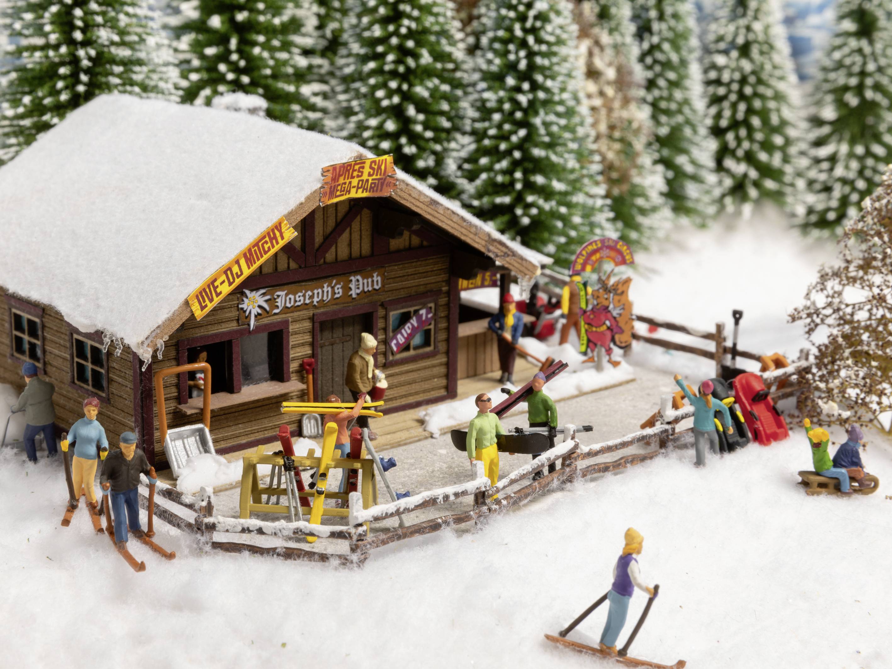 Een besneeuwde miniatuurscène toont Joseph's Pub met skiërs en sneeuwpoppen. Mensen zijn bezig in winterkleding.
