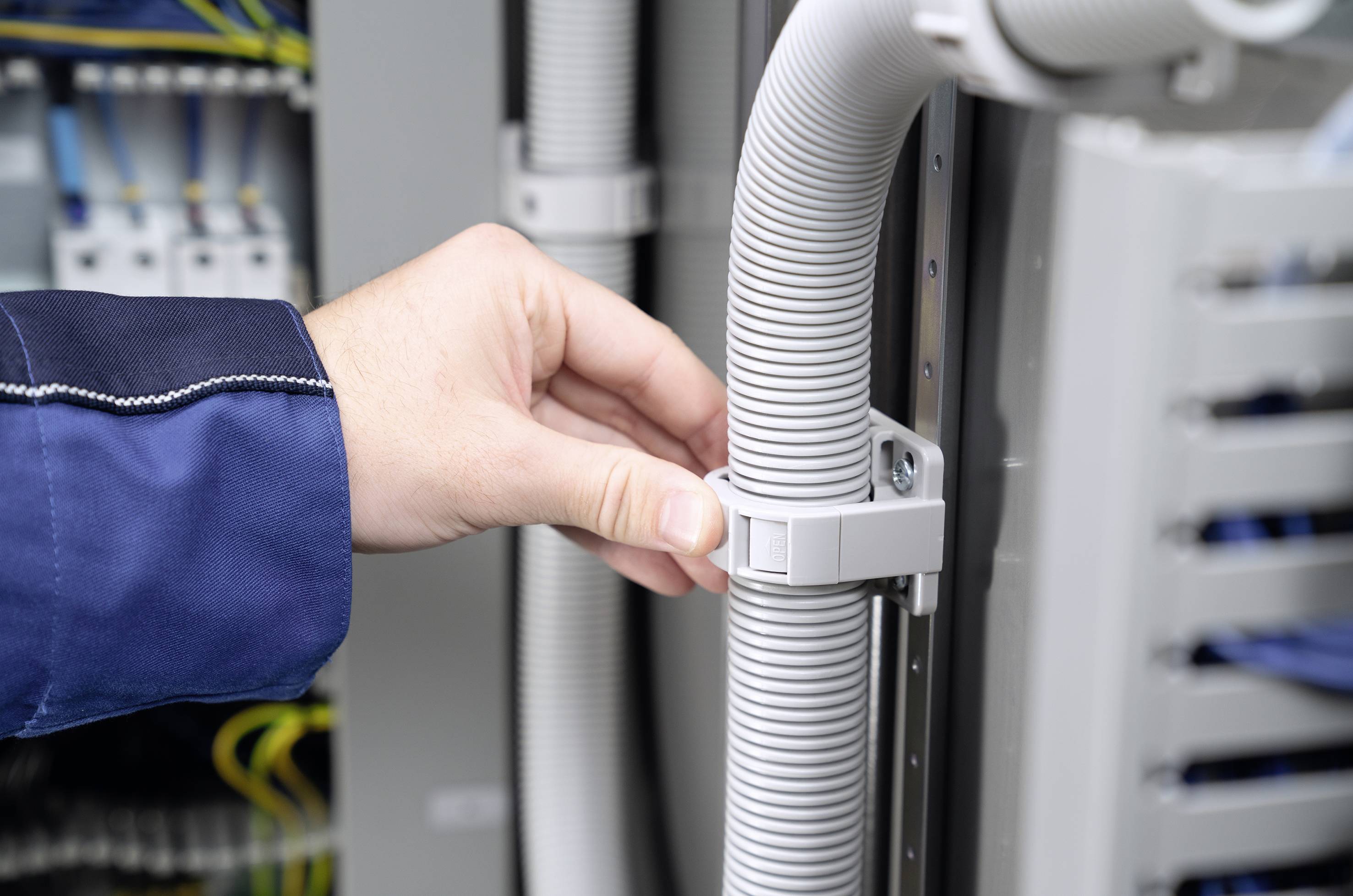 Een hand die een grijs kabel vast houdt in een zekeringkast. Op de achtergrond zijn nog meer kabels en aansluitingen zichtbaar.