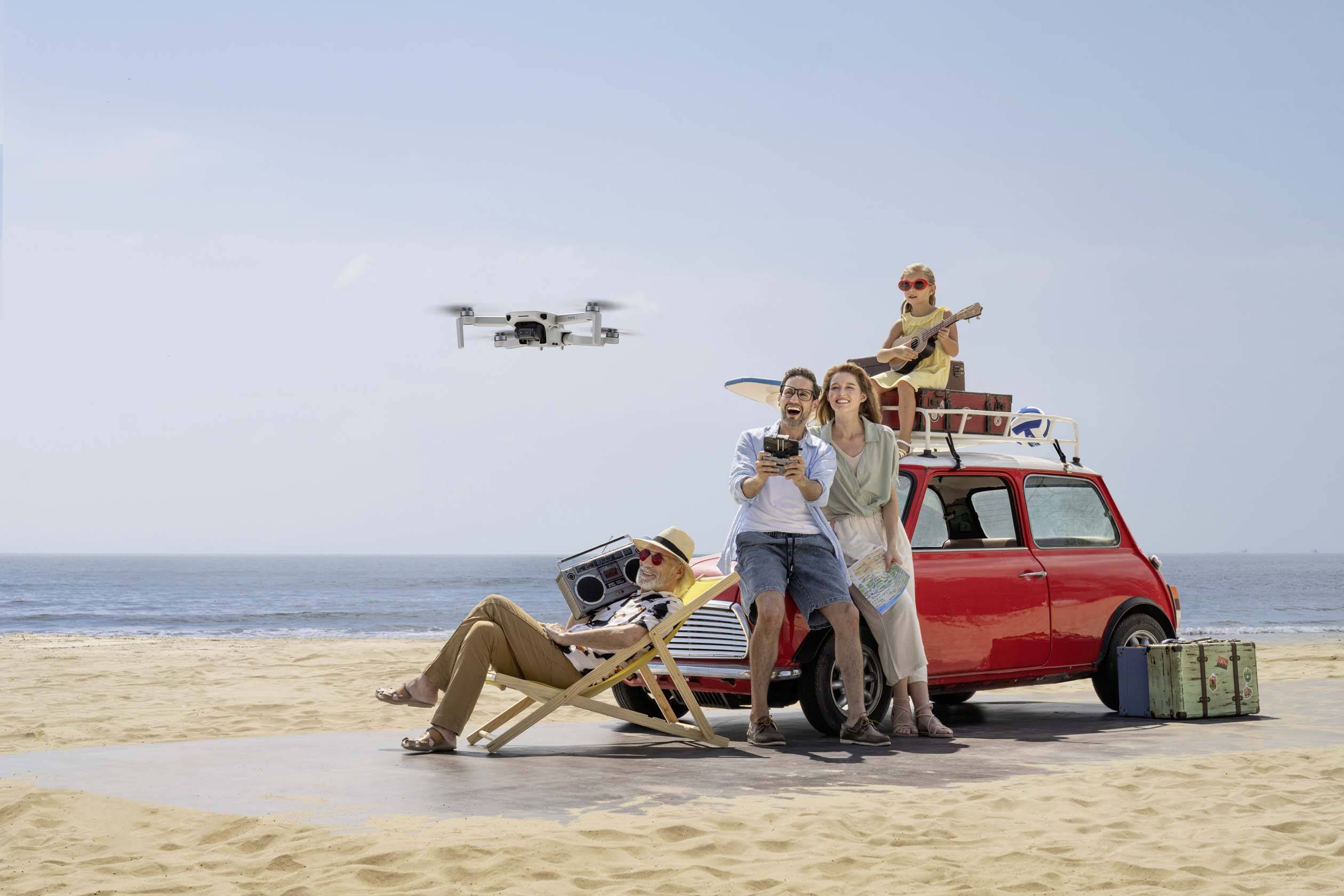 Een groep van vier personen ontspant zich op het strand naast een rode auto. Een man bestuurt een drone, terwijl anderen zich ontspannen.