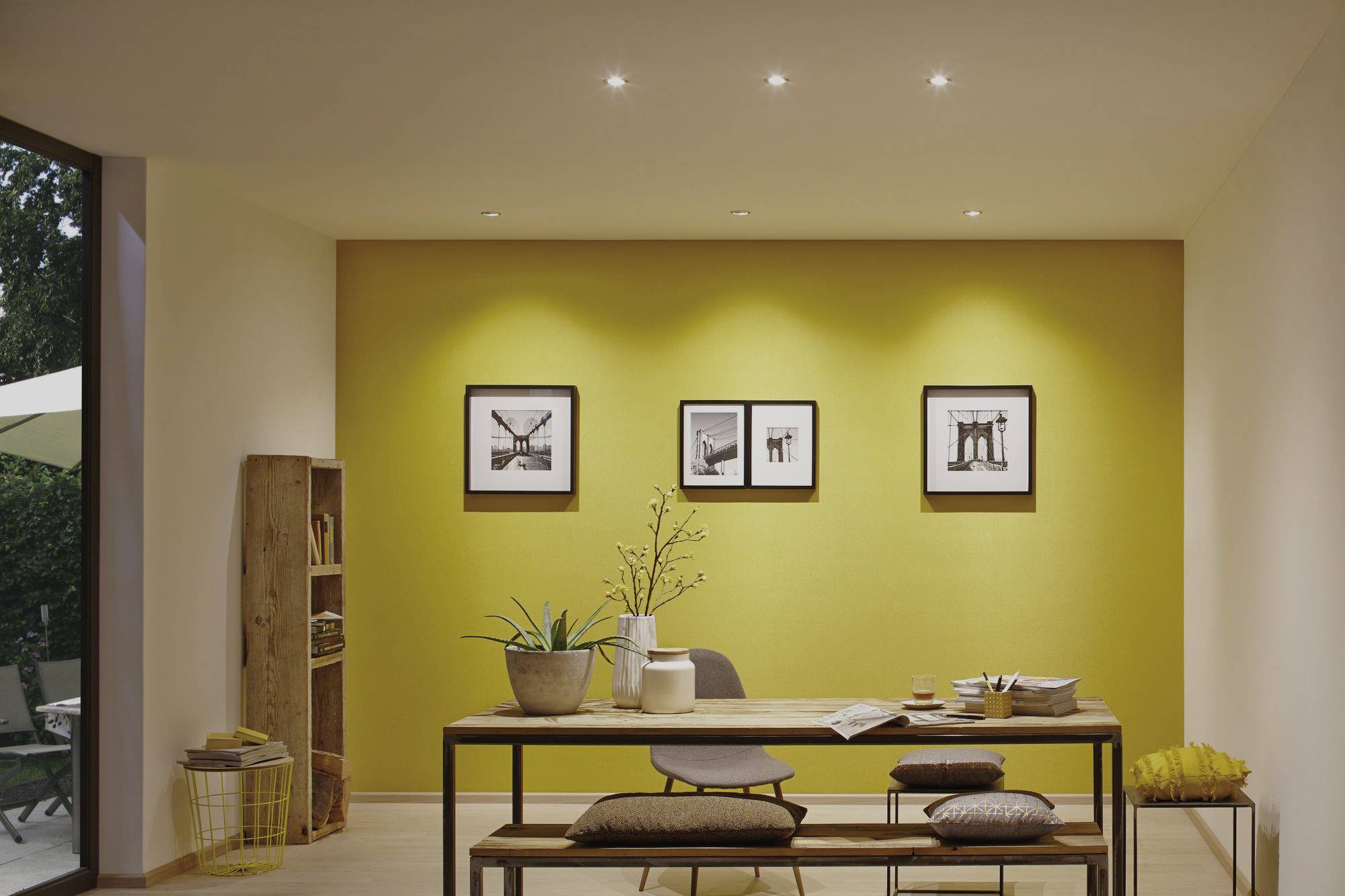Moderne eetkamer met gele accentmuur, houten bank en tafel. Drie zwart-wit foto's aan de muur. Helder licht van bovenaf.
