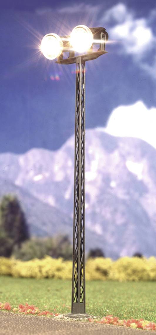 Een hoge straatlantaarn met twee felle lampen staat voor een bergachtergrond. Lichte wolken en bomen op de achtergrond.