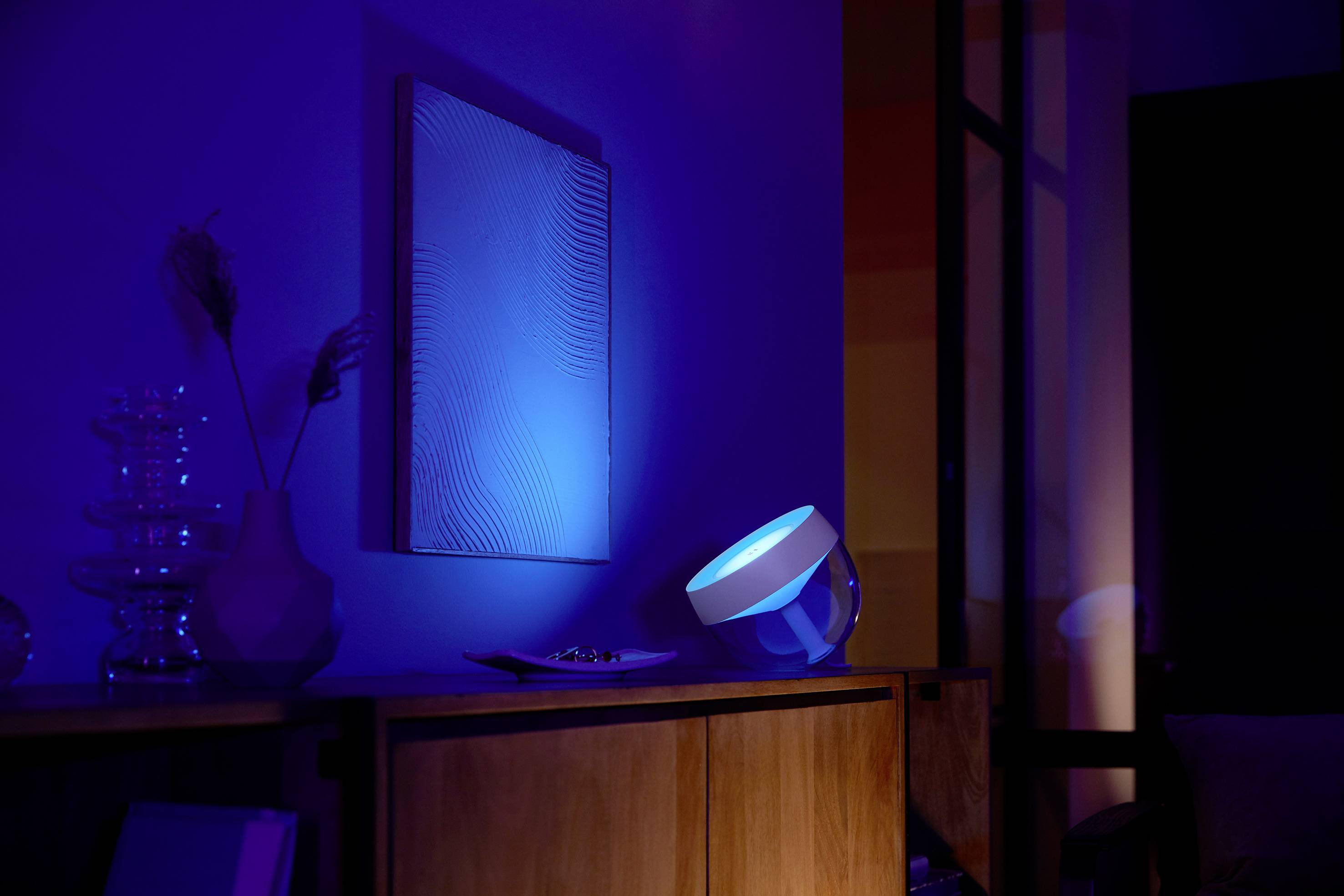 Een woonkamer met dimbaar licht, een blauw licht schijnt op een kunstwerk aan de muur. De ruimte oogt modern en sfeervol.