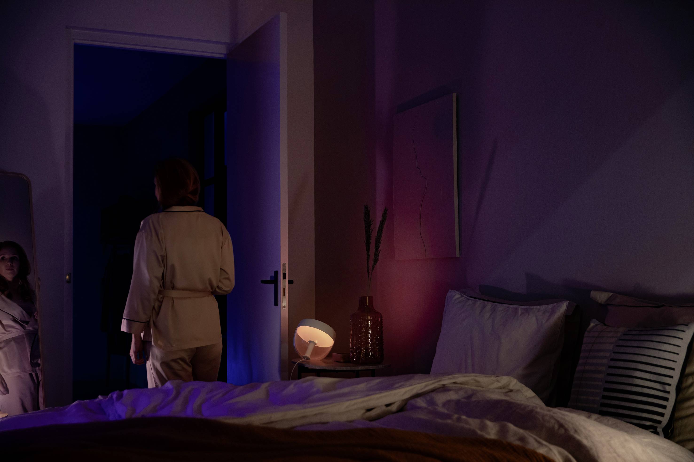 Een persoon in een pyjama staat in een spaarzaam verlichte slaapkamer naast een open deur, het bed is onaangemaakt.