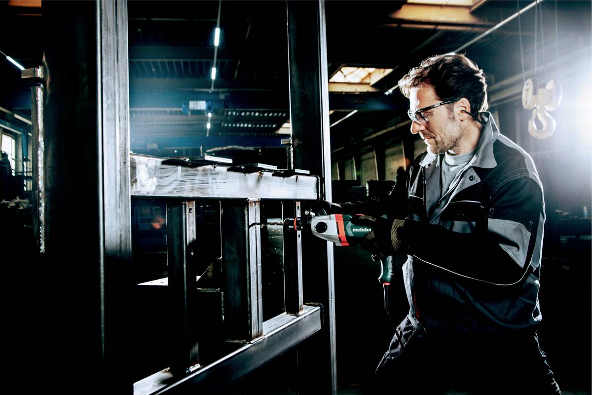 Een arbeider in werkkleding gebruikt een elektrische handboormachine in een fabriekromgeving om een metalen onderdeel te boren.