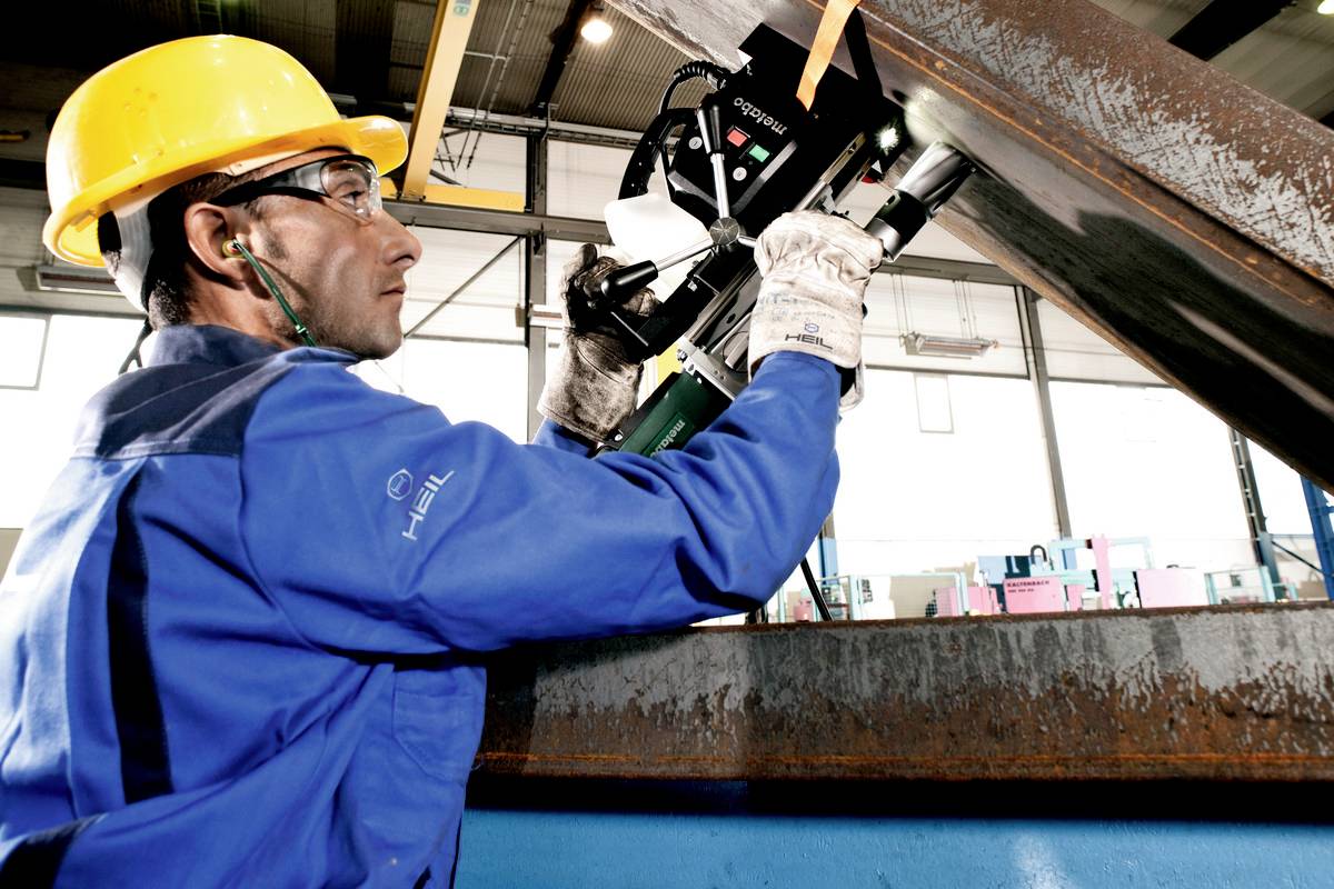 Een arbeider in beschermende kleding en helm bedient een handmatig snijgereedschap in een fabriekshal. De omgeving is industrieel.