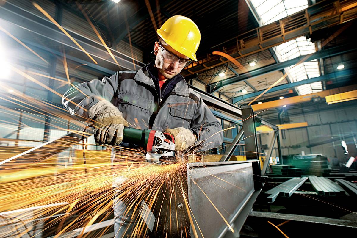 Een arbeider in beschermende kleding slijpt metaal met een haakse slijper, waarbij vonken in de industriële omgeving spatten.