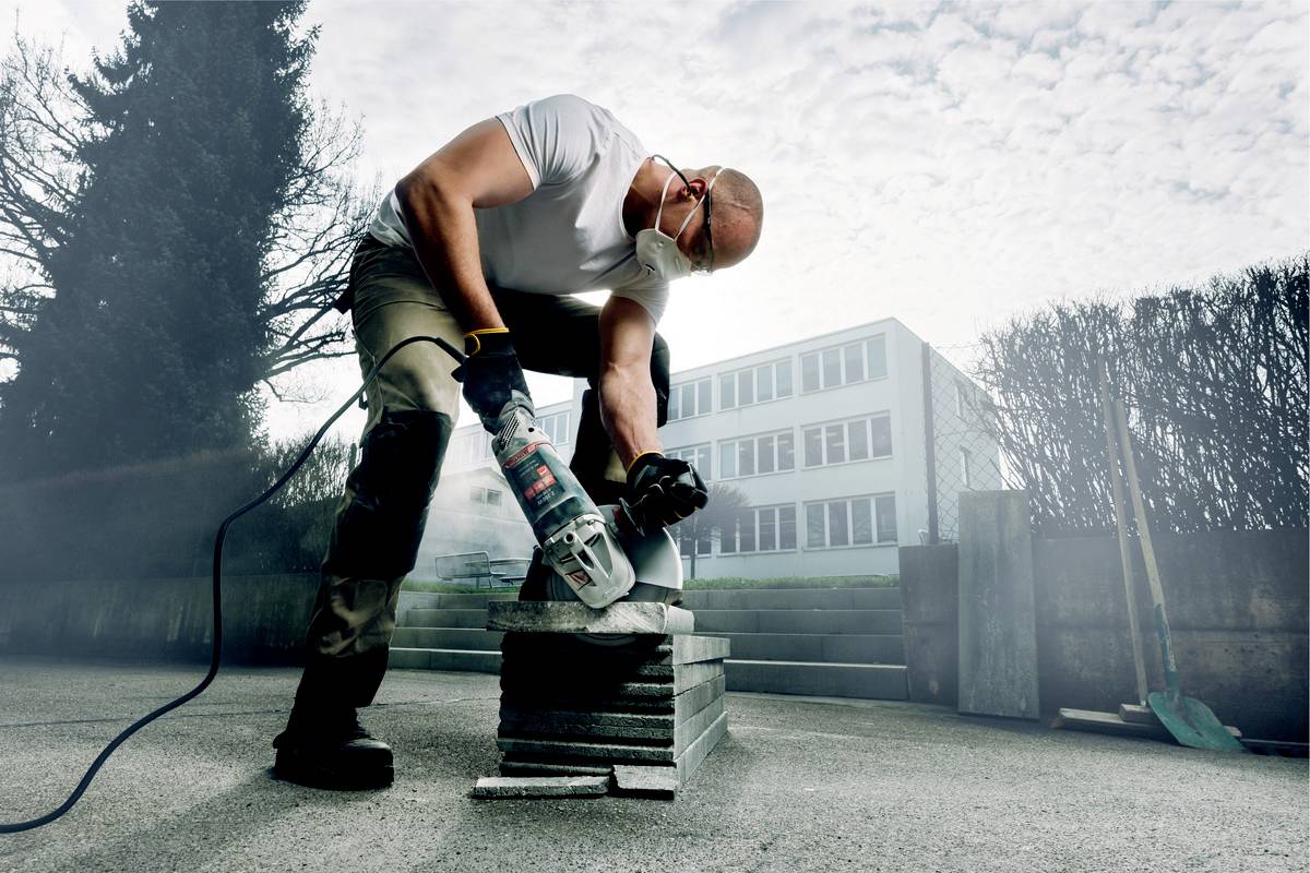 Een man draagt beschermende kleding en snijdt betonplaten buiten met een elektrische zaag. Op de achtergrond is een gebouw zichtbaar.
