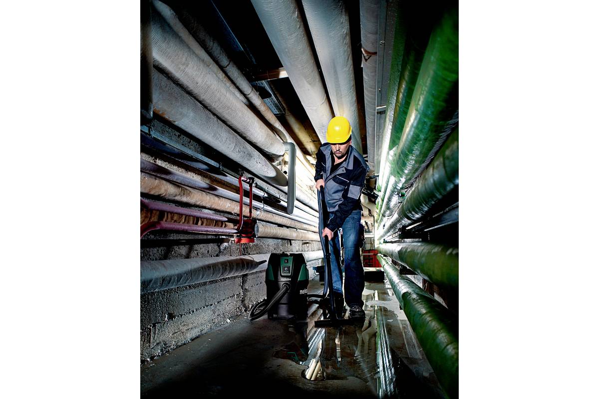 Een arbeider met een gele helm reinigt een smal industrieel gangpad met buizen aan weerszijden. Op de vloer zijn plassen water aanwezig.