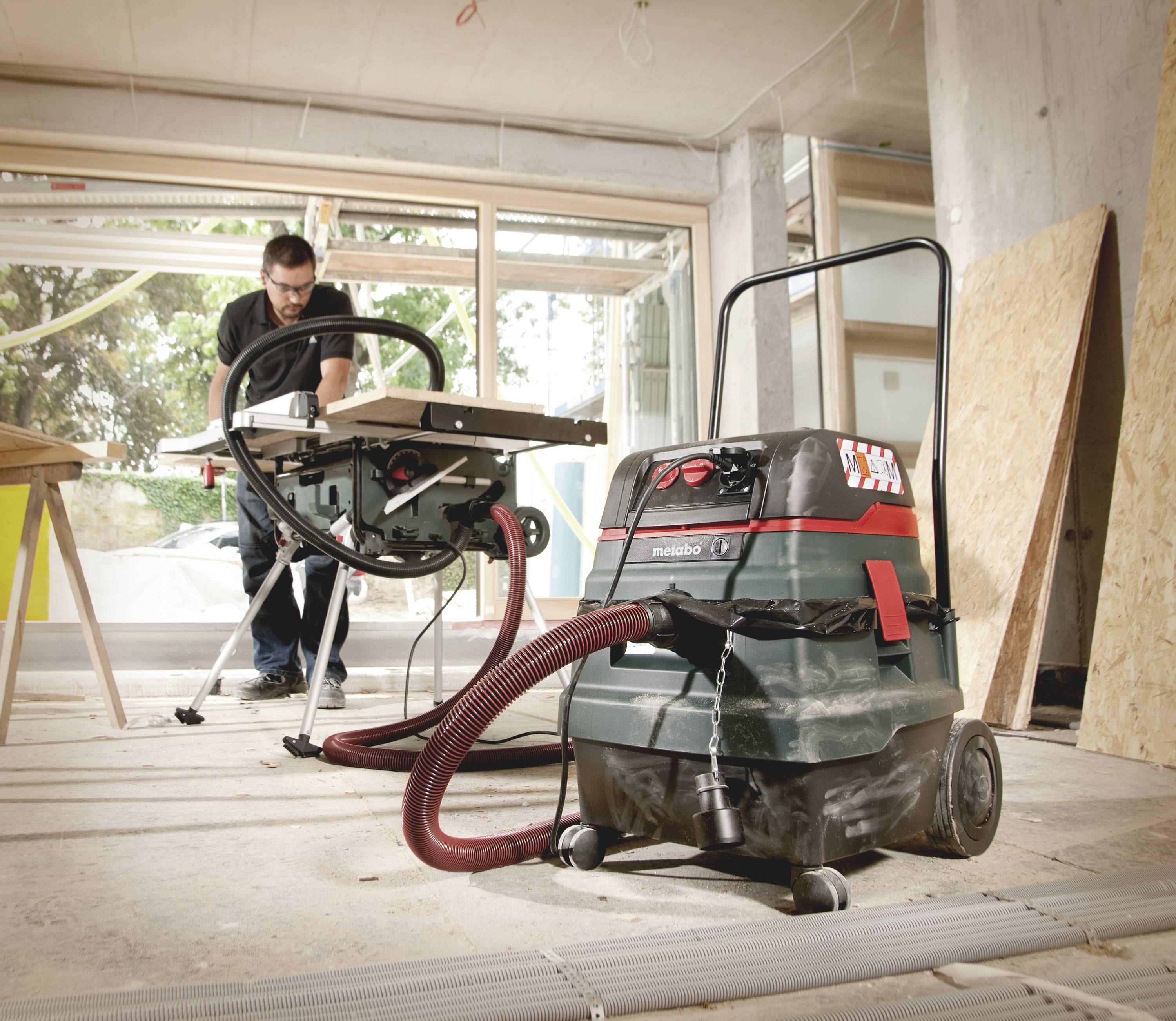 Een man bedient een tafelzaag in een werkplaats, met een vacuümsysteem aangesloten voor stofafzuiging. Houten panelen en bouwmaterialen zijn zichtbaar.