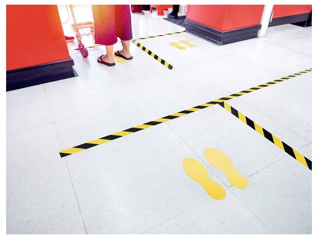 Een persoon staat op veilige afstand in de rij, gemarkeerd door gele voetafdrukken en zwart-gele gestreepte tape op de vloer.