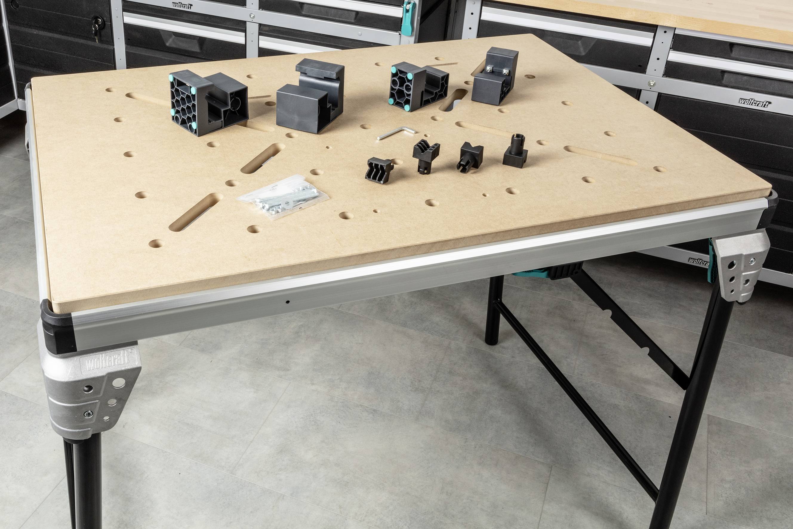 Werktafel met geperforeerd oppervlak, waarop verschillende zwarte kunststofonderdelen, schroeven en twee zilveren blokken zijn gerangschikt.