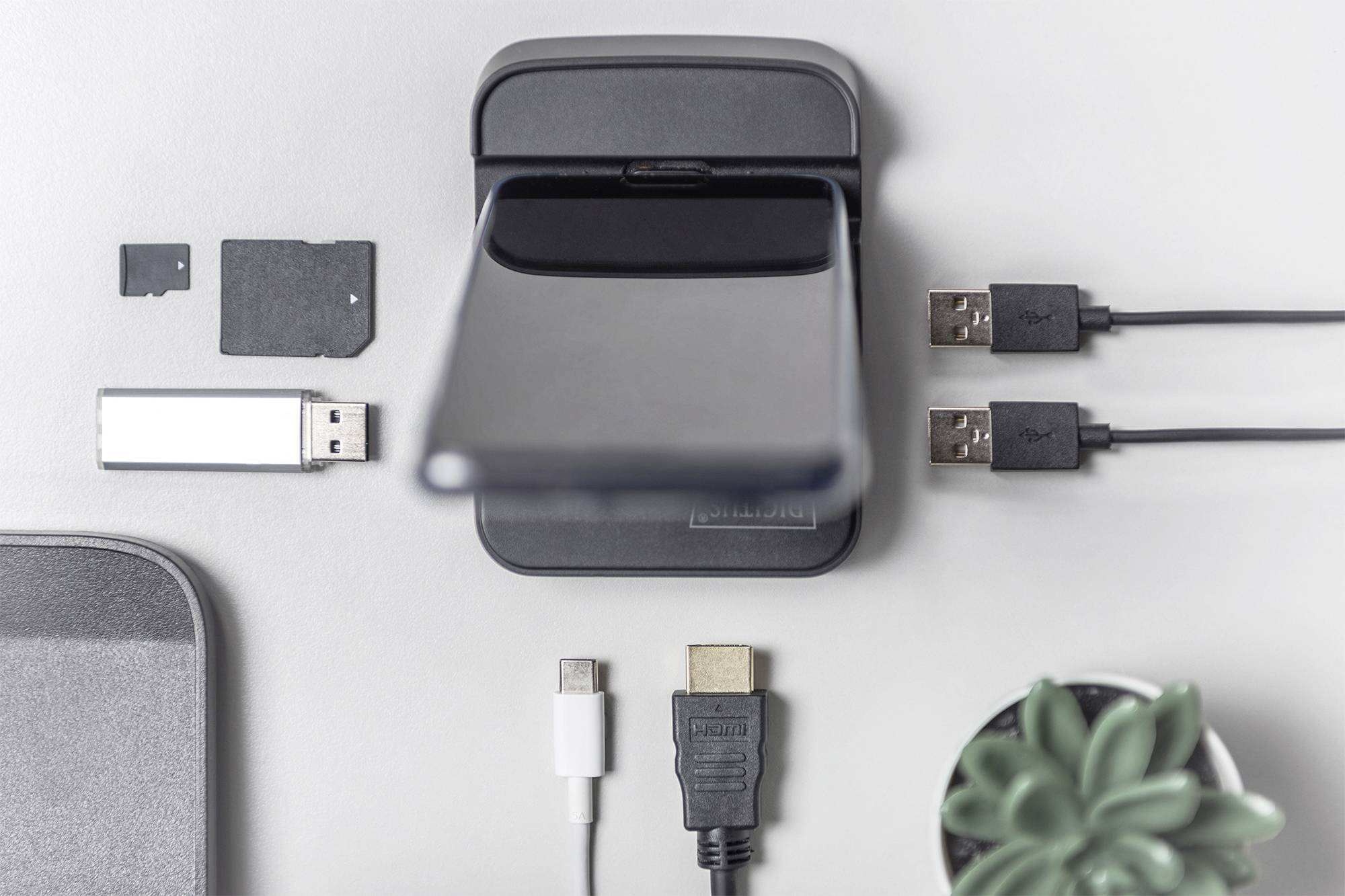 Smartphone op draadloos laadstation, omringd door USB-sticks, SD-kaarten en kabels op een witte tafel. Rechtsonder een kleine plant.