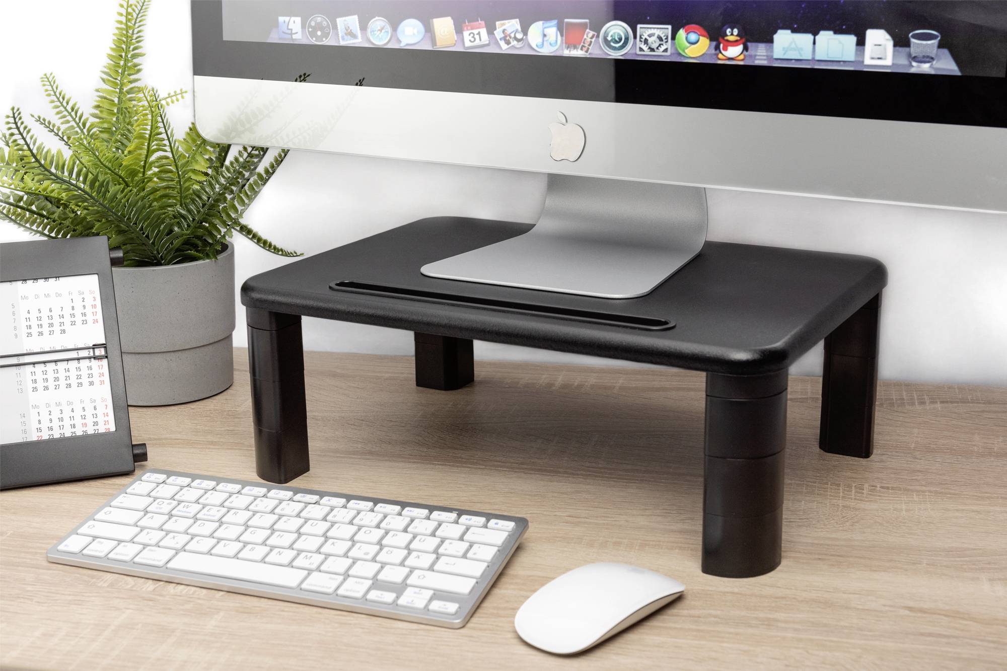 Een bureau met een computerscherm op een monitorstandaard, ernaast een toetsenbord, een muis en een plant.