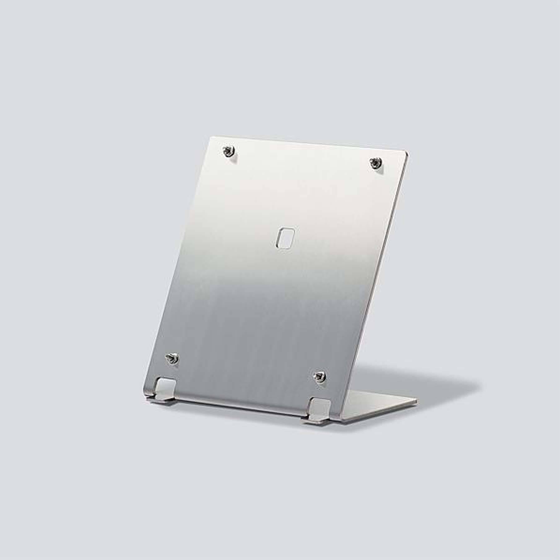 Een zilveren, rechthoekige metalen standaard met vier zichtbare schroeven, staat op een lichtgrijze oppervlakte.
