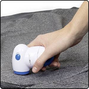 Hand houdt een pluizenverwijderaar boven een grijze stof om pluisjes te verwijderen. Apparaat is wit met een blauw detail.