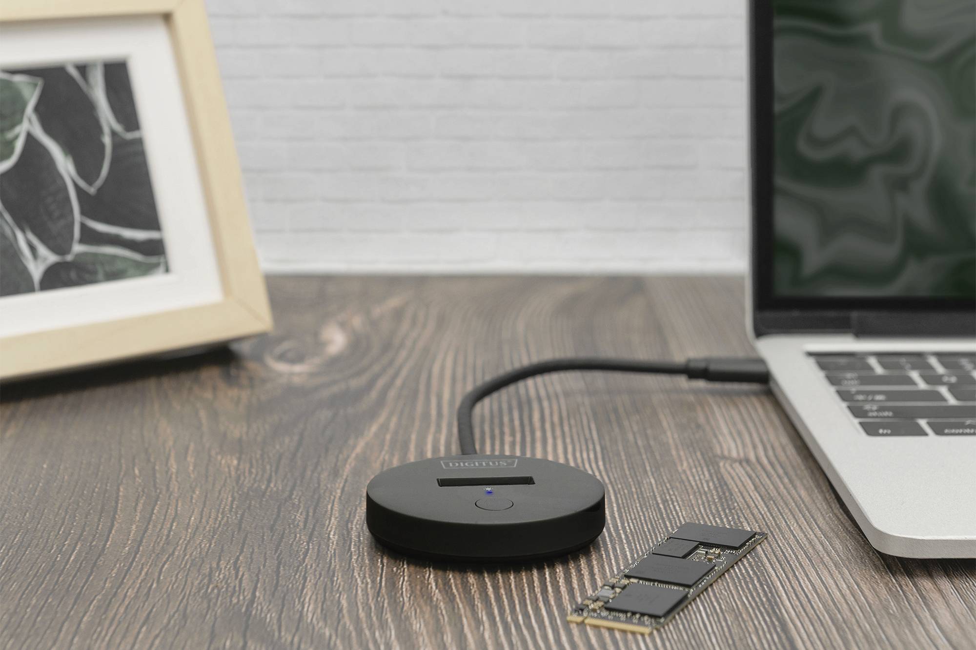 'Een ronde adapter met kabel verbindt een laptop op een houten tafel. Een fotolijst staat op de achtergrond, ernaast ligt een geheugenmodule.'