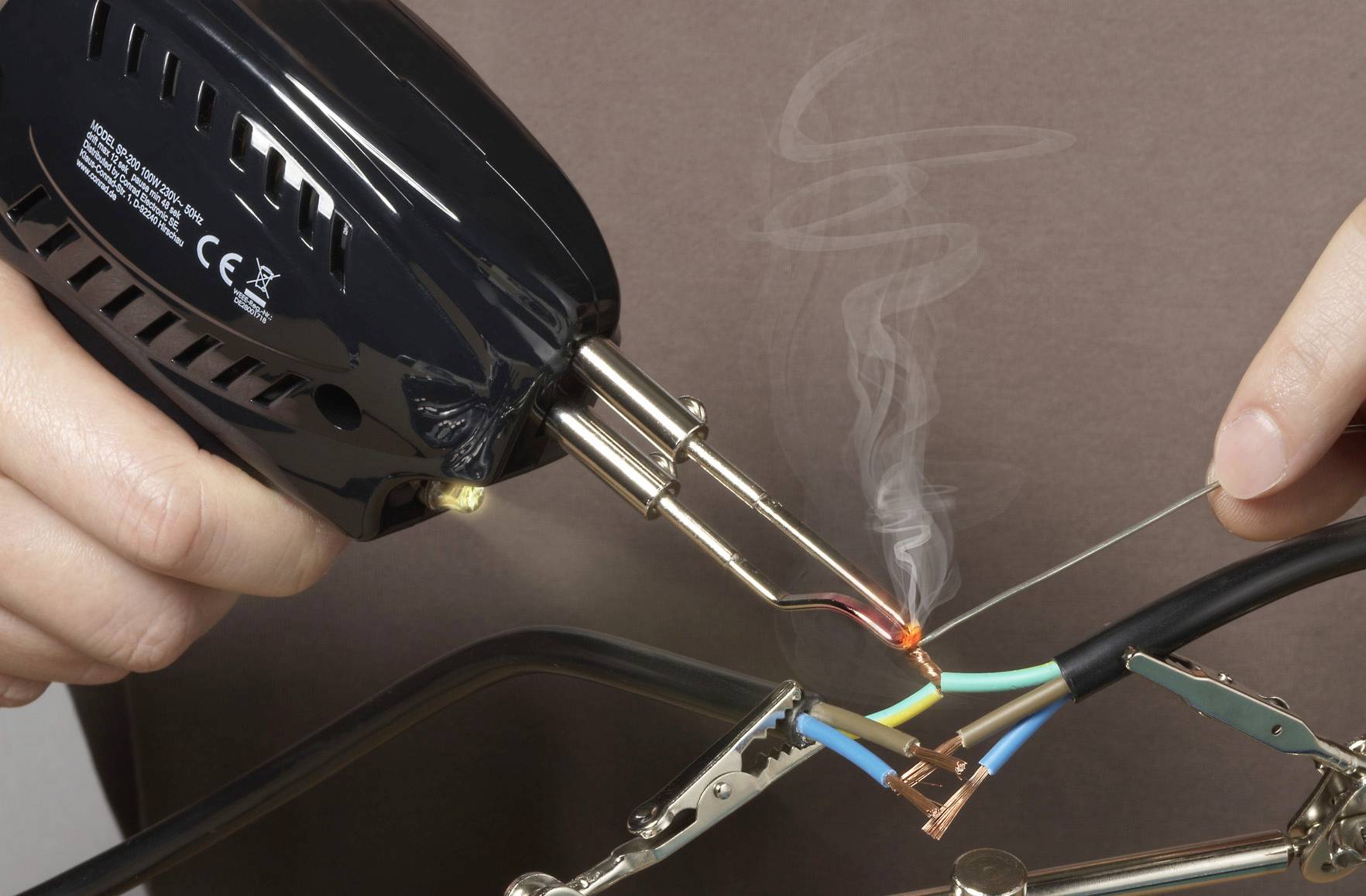 'Een persoon soldeerd twee draden. Rook stijgt op van de soldeerplaats. De draden zijn herkenbaar door gekleurde isolatielagen.'