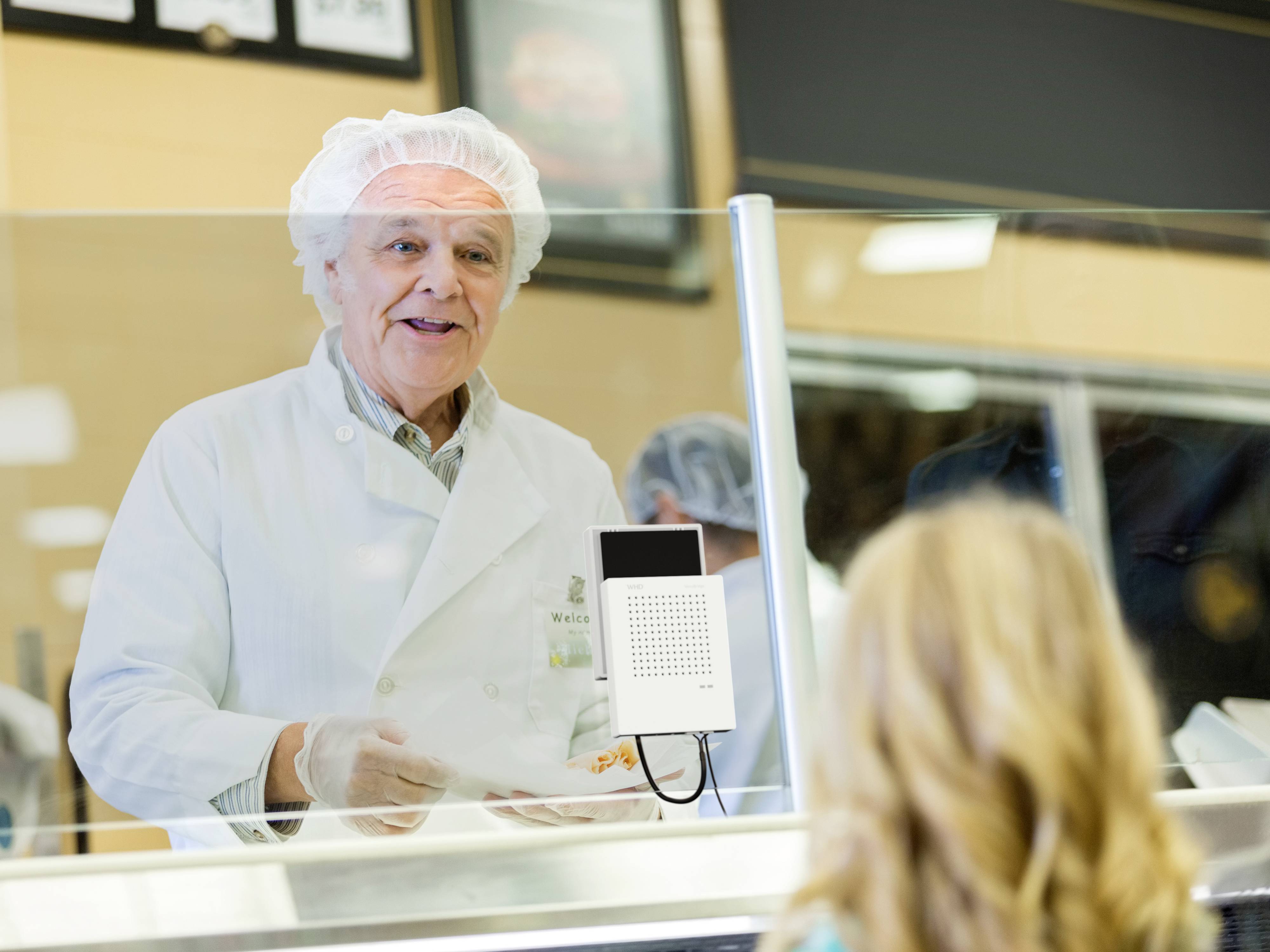 Een oudere man in een witte laboratoriumjas en met een haarnet bedient een klant in een delicatessenzaak.