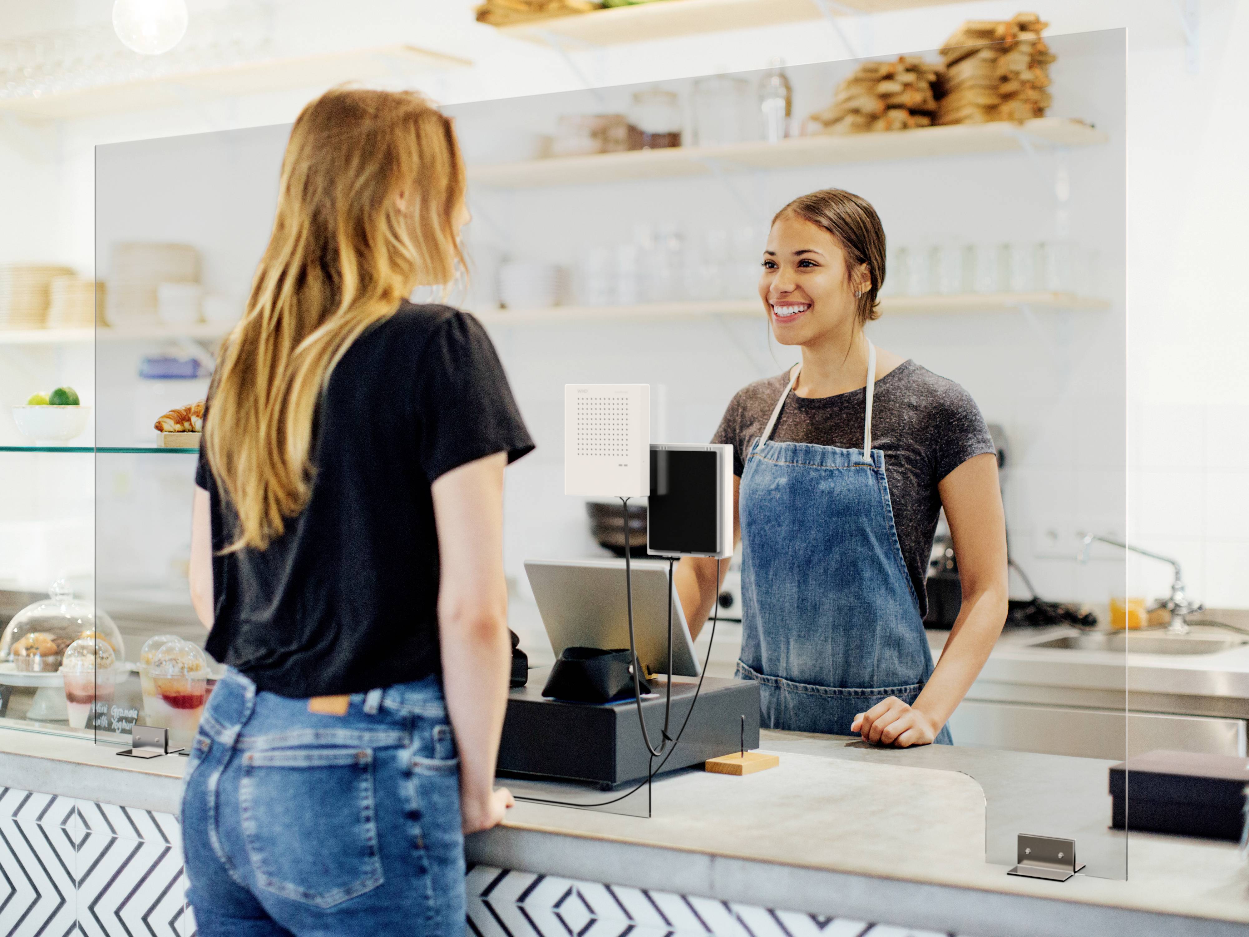 Klant staat bij de toonbank van een café en spreekt met een glimlachende verkoopster. Beiden worden gescheiden door een plexiglazen scherm.
