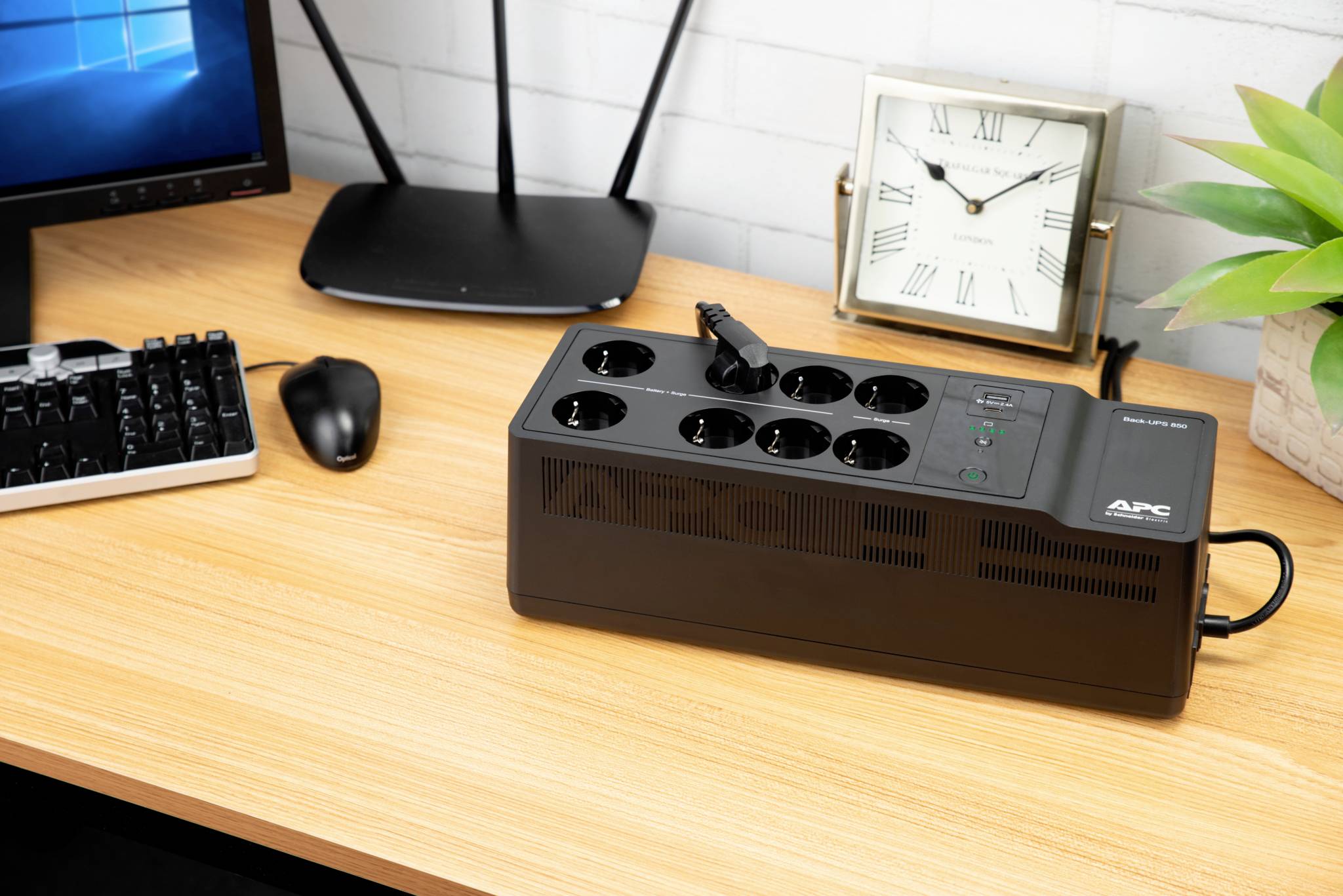 Op een bureau staat een APC-overspanningsbeveiliging met meerdere stopcontacten. Ernaast bevinden zich een monitor, een toetsenbord, een muis en een klok.