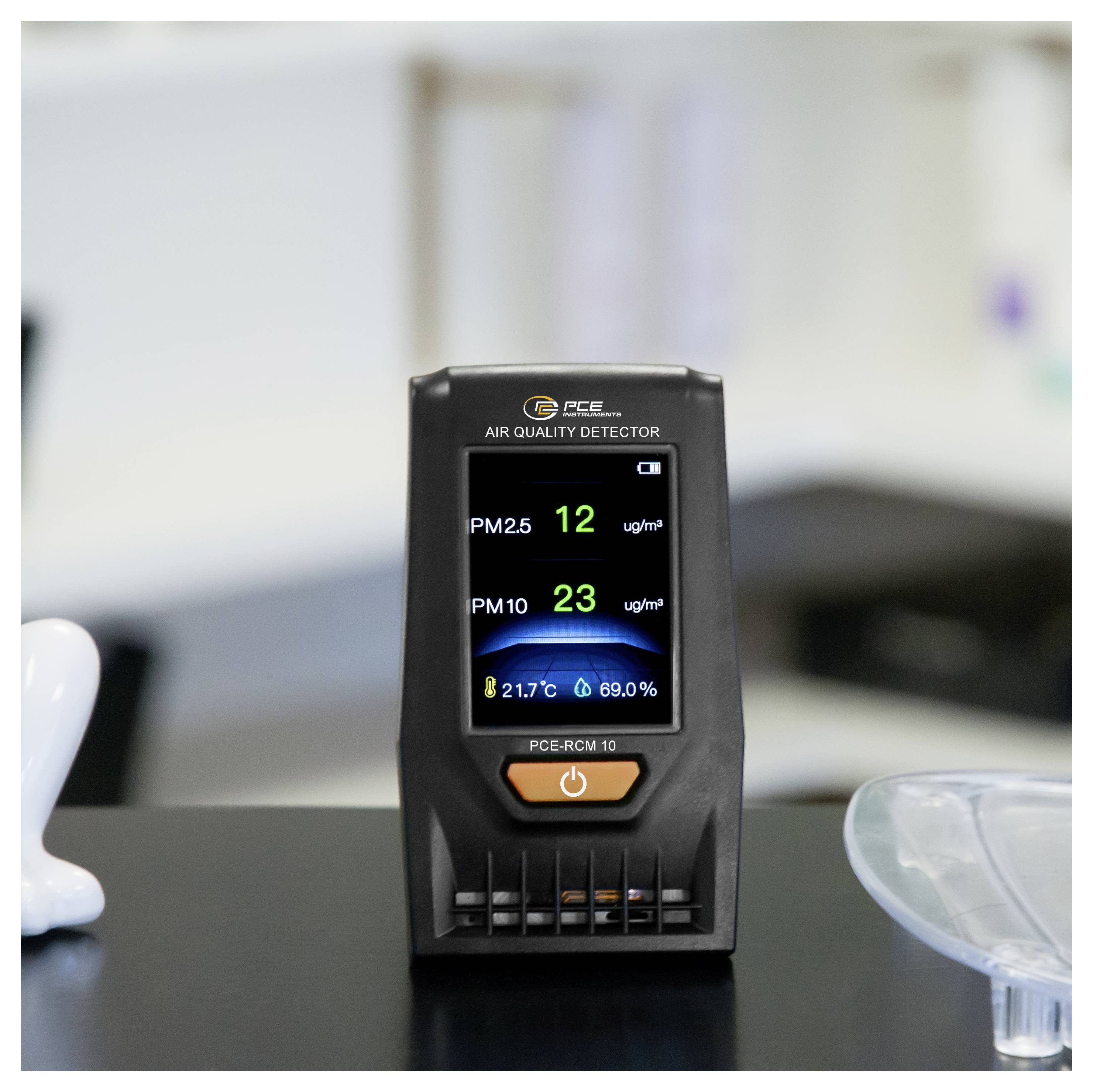 Draagbare luchtkwaliteitsmeter op een bureau met PM2,5 12 μg/m³, PM10 23 μg/m³, 21,7°C en 69,0% luchtvochtigheid.