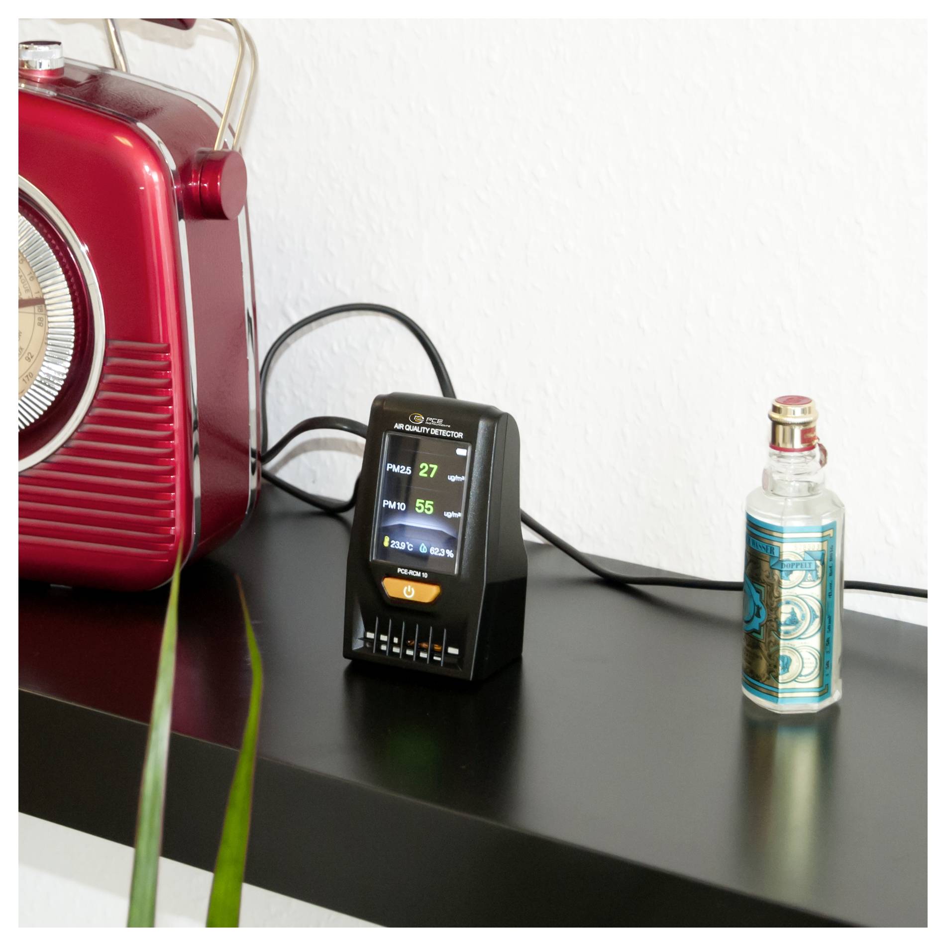 Een kleine luchtkwaliteitsmonitor op een zwarte plank geeft PM2,5 27 en PM10 55 weer, naast een rode radio en een verdampingsapparaat.