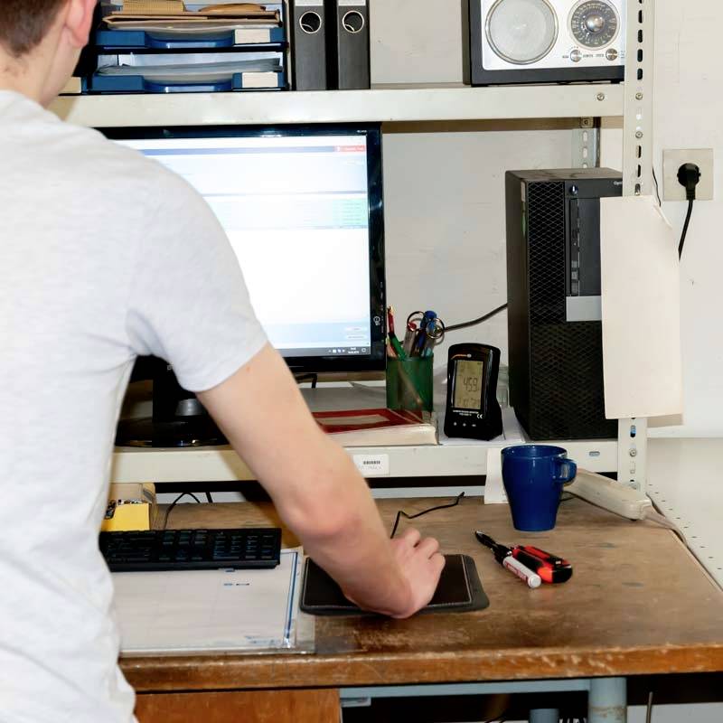 Een persoon staat bij een bureau met een computer waarop een tabel wordt weergegeven. Naast de monitor liggen kantoorartikelen.