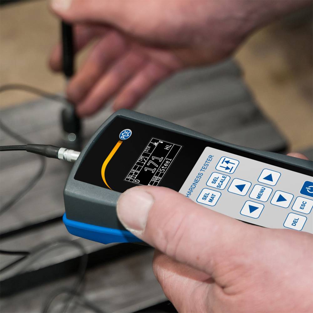Een persoon houdt een hardheidsmeetapparaat met digitaal display en toetsen, terwijl het meetapparaat is aangesloten op een metalen oppervlak.