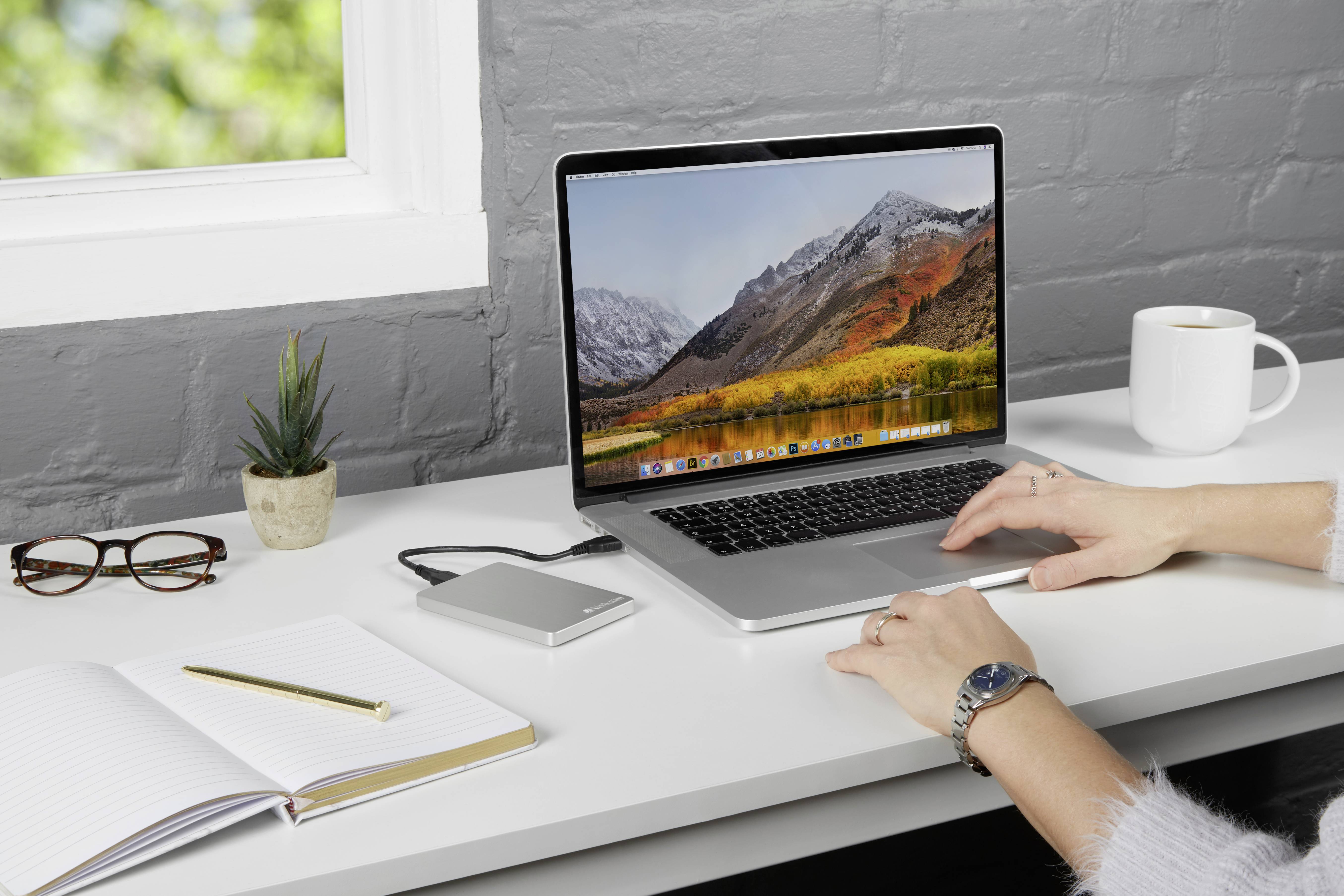 Een persoon werkt op een laptop aan een bureau met een notitieboek, bril, externe harde schijf, plant en koffiekopje.