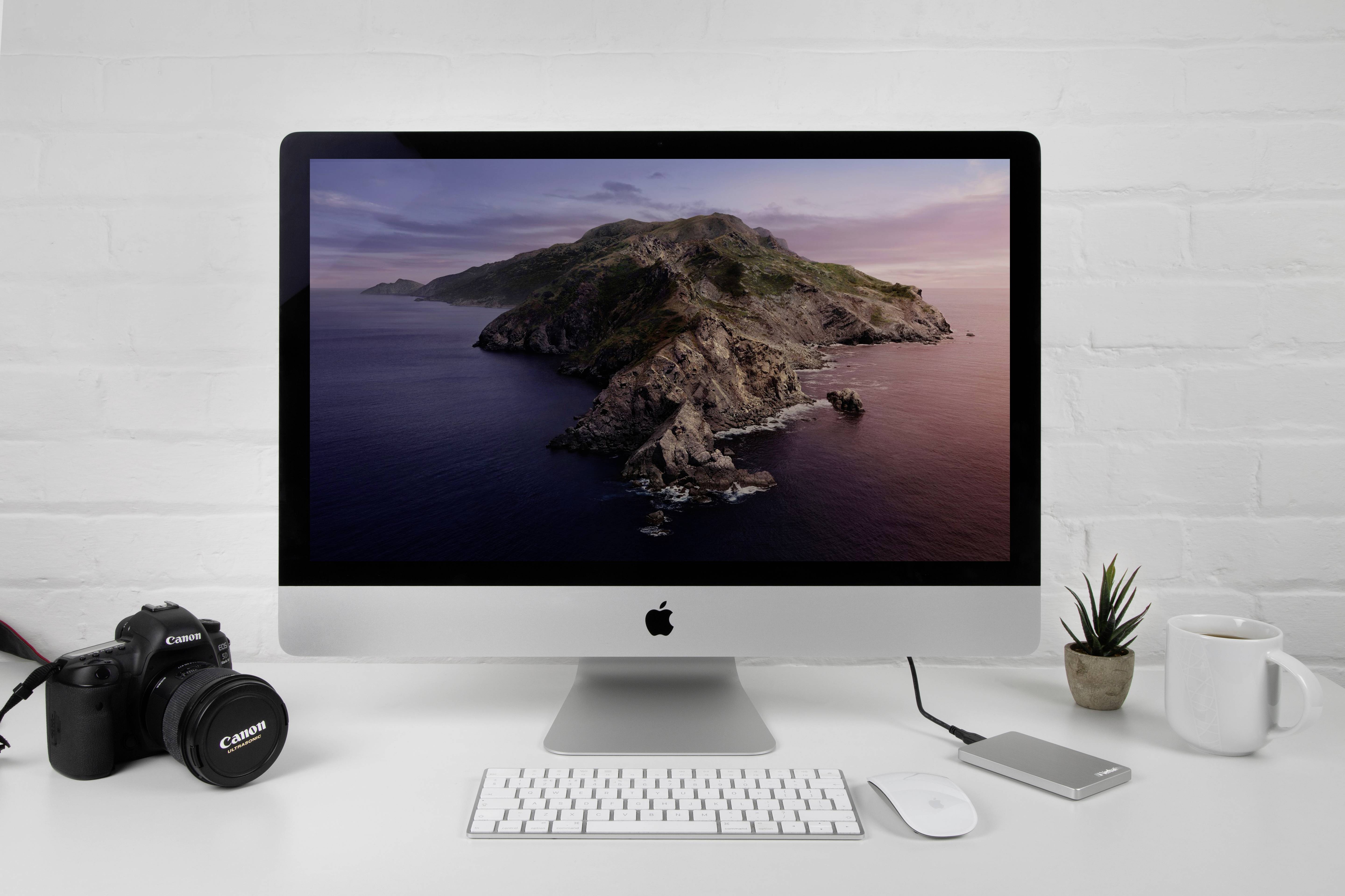 Een bureau met een Apple-computer waarop een kustlandschap wordt weergegeven, ernaast een camera, een externe harde schijf en een mok.