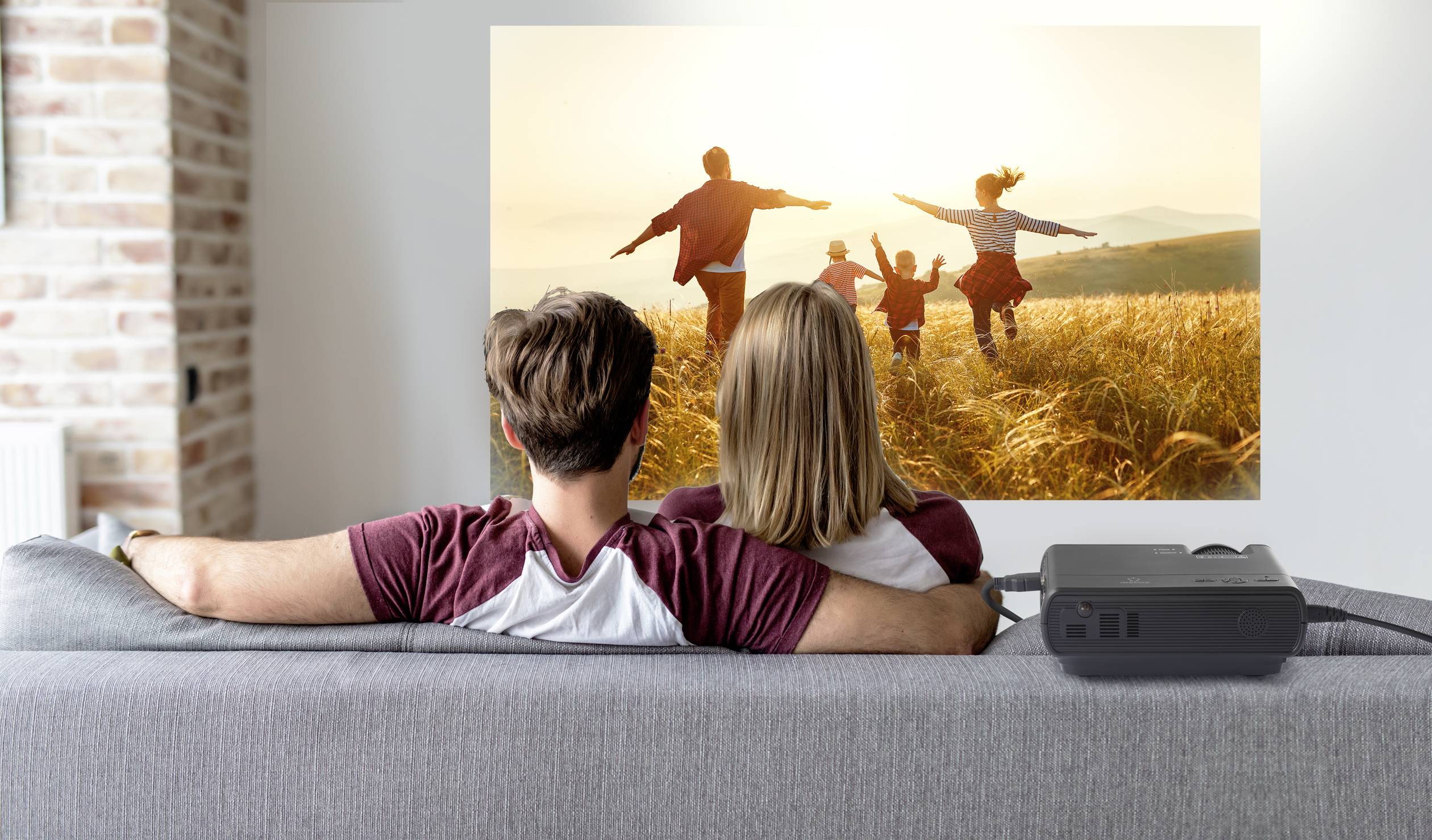 Een stel zit op een bank en kijkt naar een film op een projector die een familie toont die gelukkig over een veld loopt.