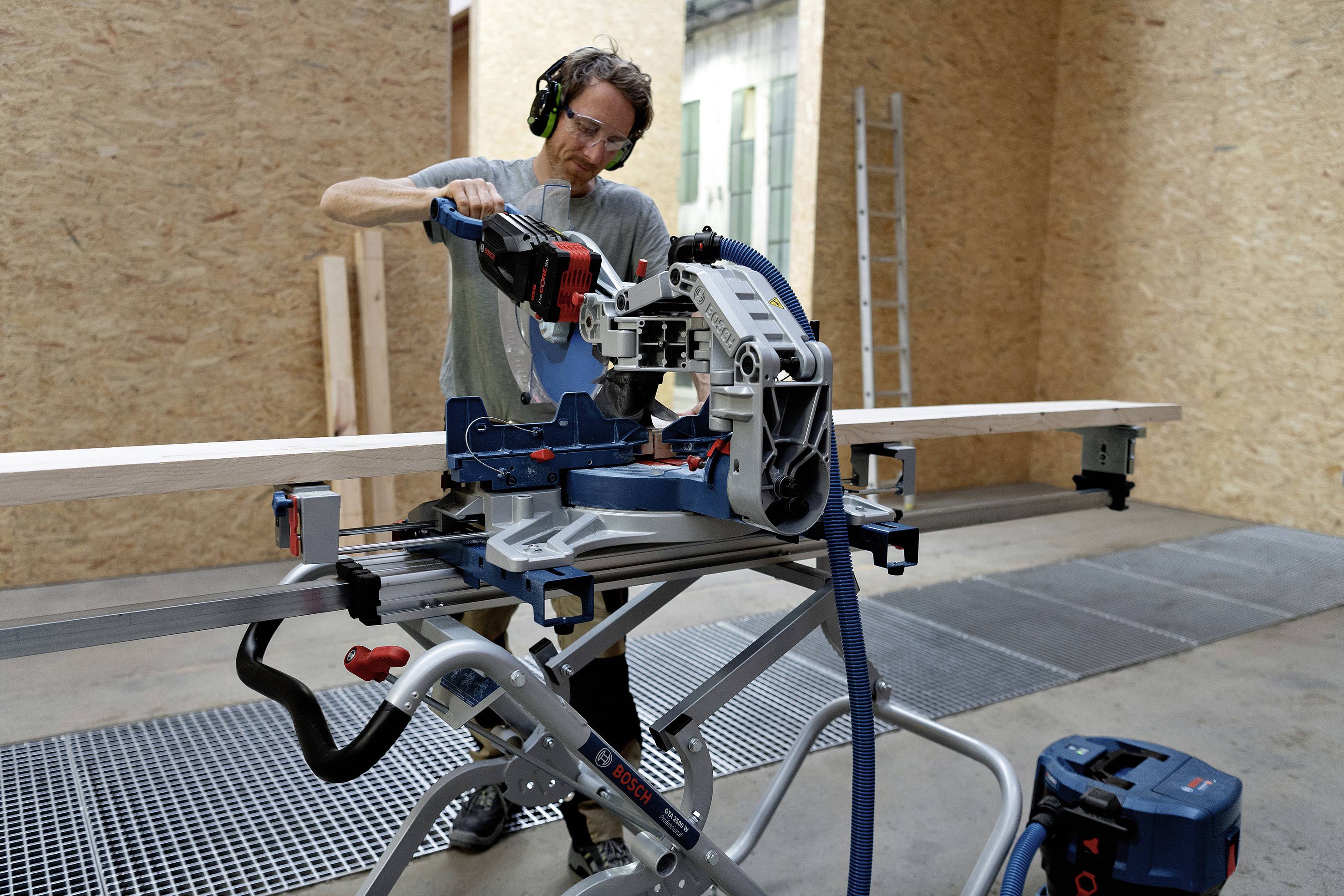Een man met gehoorbescherming zaagt hout met een elektrische verstekzaag in een werkplaats. Op de achtergrond staat een ladder.