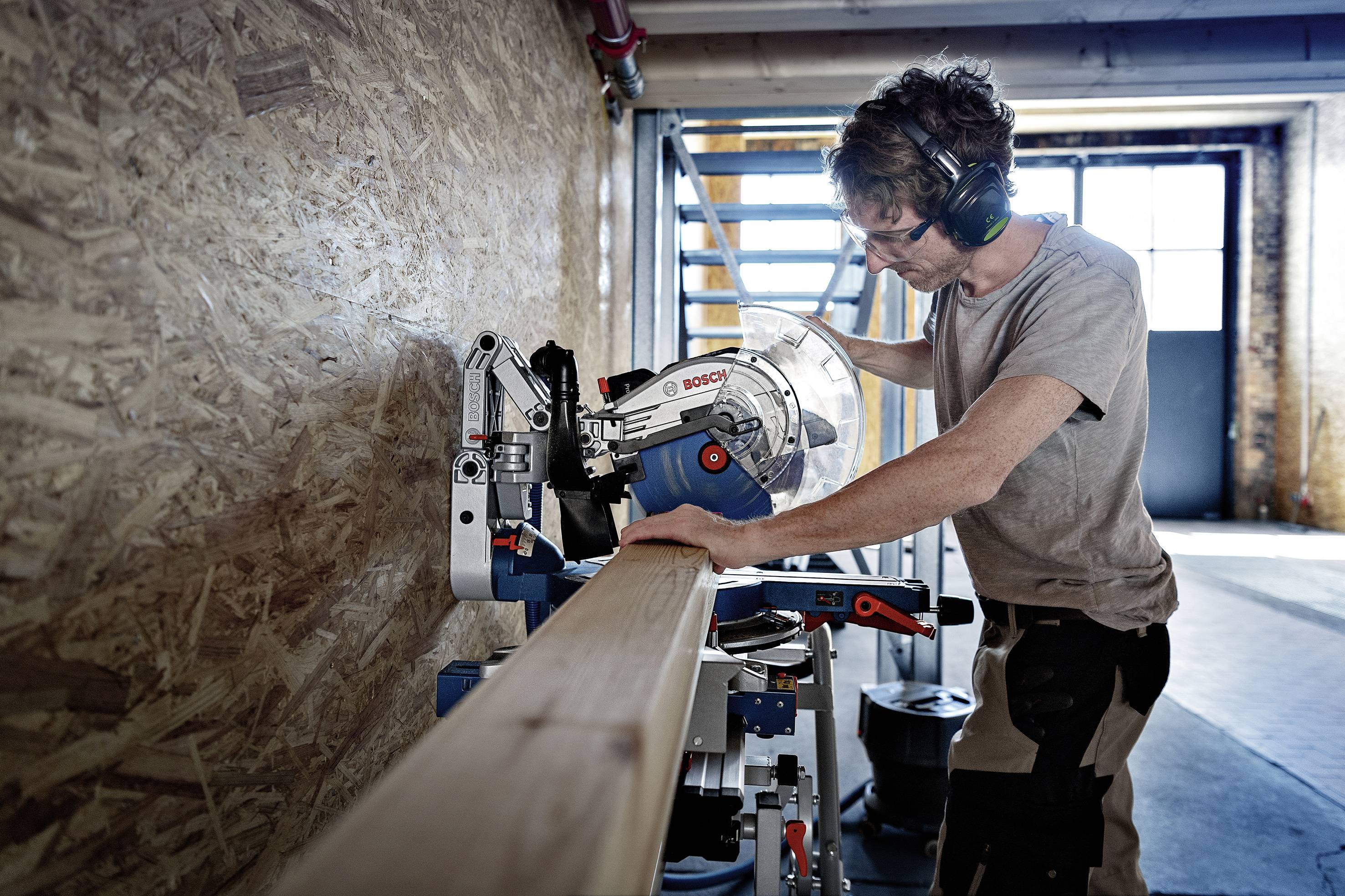 Een persoon in een werkruimte zaagt een houten plank met een elektrische zaag. Ze draagt een veiligheidsbril en gehoorbescherming.
