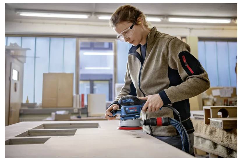 Een persoon draagt veiligheidsbril en gebruikt een elektrische schuurmachine op een houten oppervlak in een werkplaats, gefocust op zijn taak.