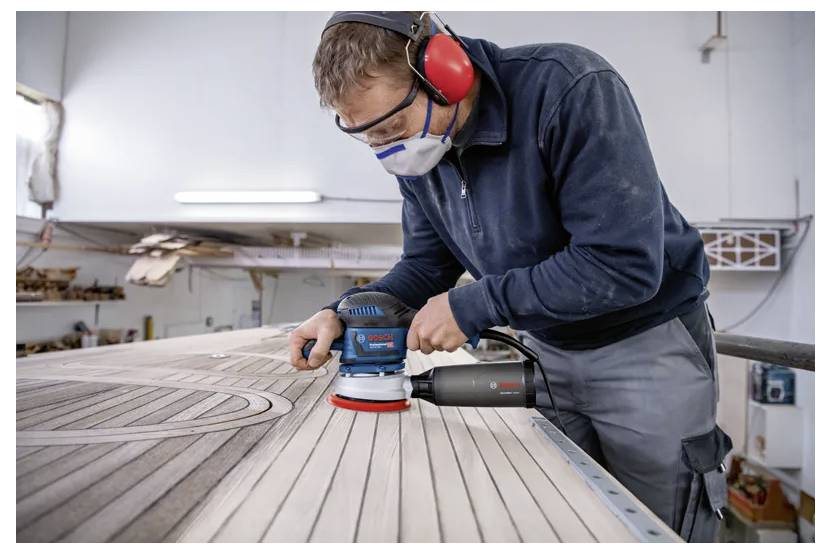 Een persoon is een groot houten oppervlak aan het schuren met een elektrische schuurmachine, gekleed in beschermende uitrusting inclusief gehoorbeschermers en een masker in een werkplaats.