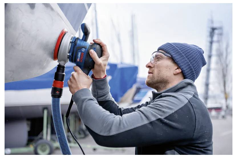 Een persoon met een muts gebruikt een elektrisch gereedschap om de romp van een boot te polijsten in een jachthaven. Vervaagde boten en masten zijn zichtbaar op de achtergrond.