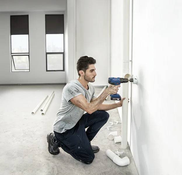 Een persoon knielt in een onafgewerkte ruimte en gebruikt een boormachine om buizen te installeren aan een witte muur. Buizen en koppelingen zijn verspreid over de vloer.