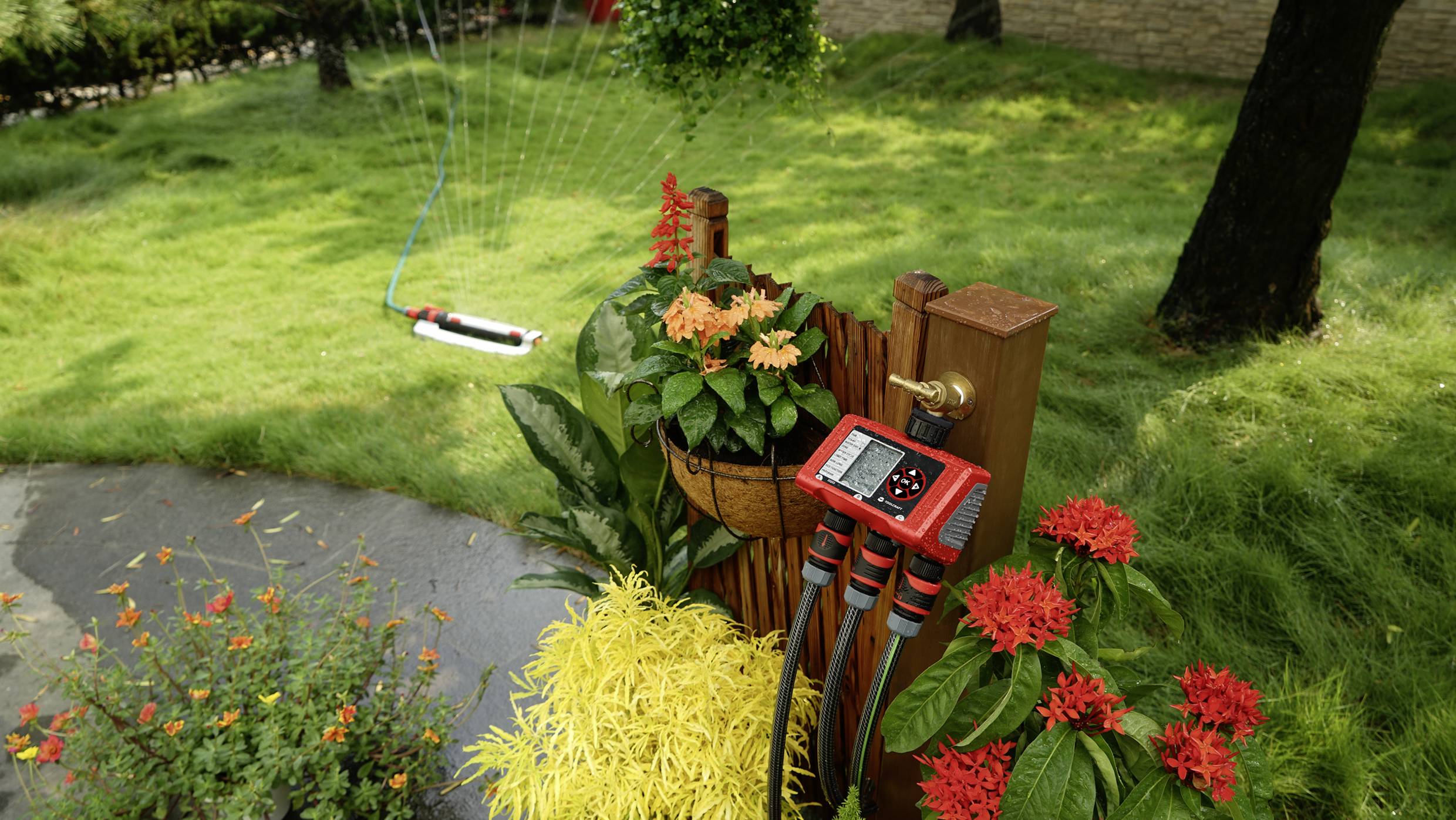 Een tuin met kleurrijke bloemen, een irrigatiesysteem met digitale besturingen en een sproeier op de achtergrond op een grasveld.