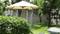 Een grote parasol werpt schaduw in een groene tuin met bomen en een ronde tafel ernaast.