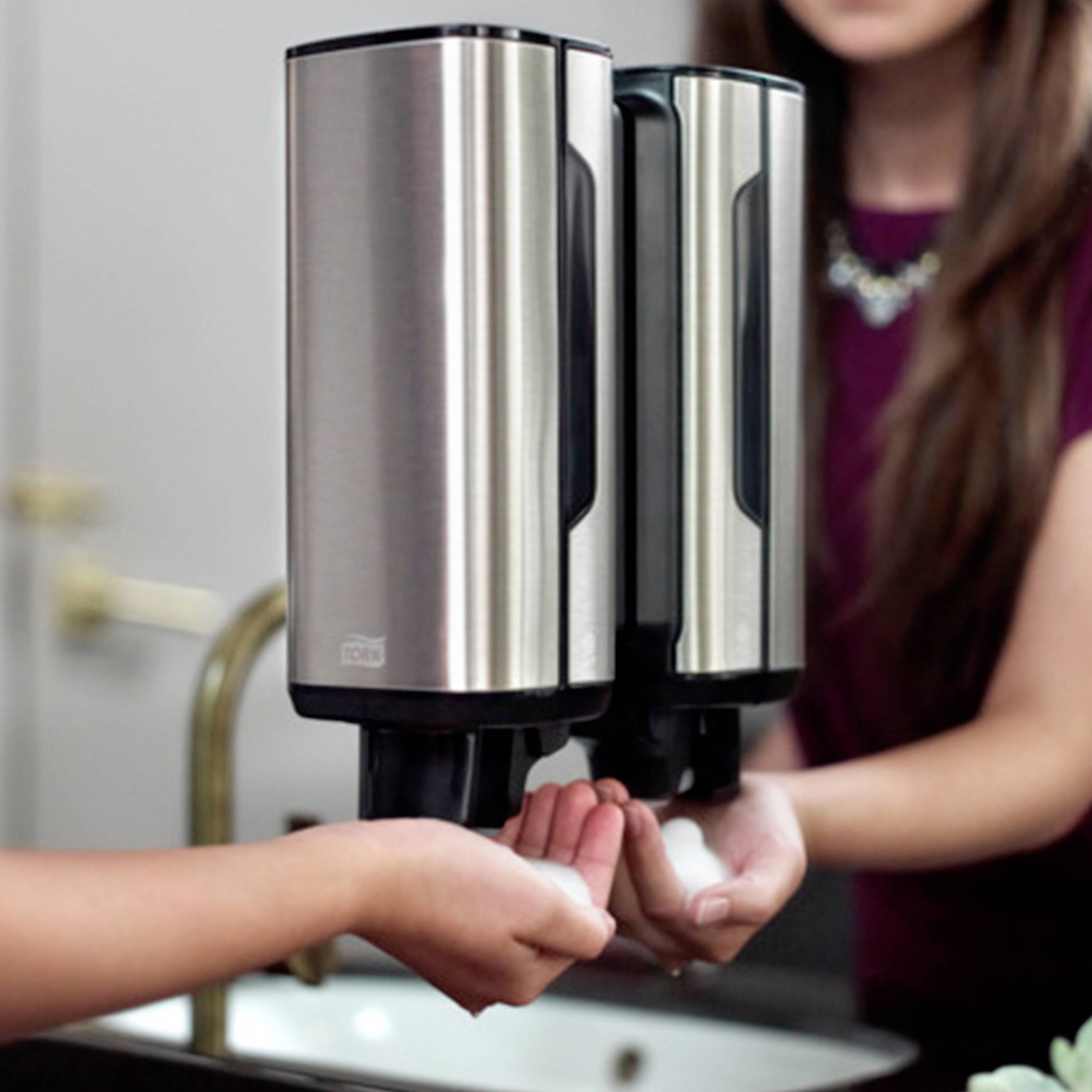 'Handen onder een zeepdispenser in de badkamer. Een persoon drukt de dispenser in, terwijl de kraan op de achtergrond zichtbaar is.'