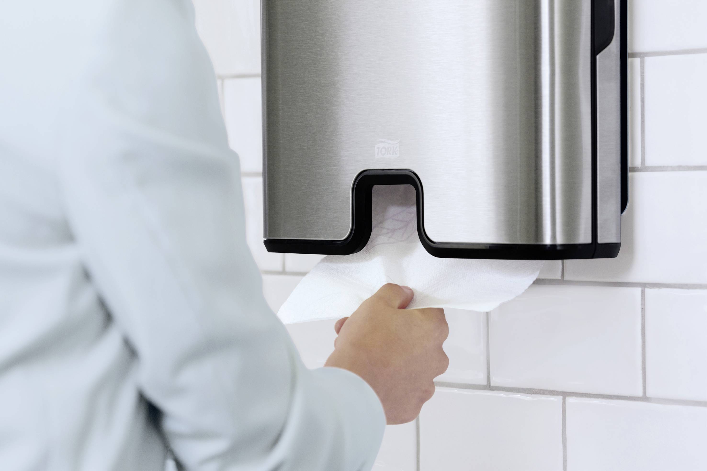 Een persoon haalt een papieren doek uit een metalen dispenser aan een betegelde muur.