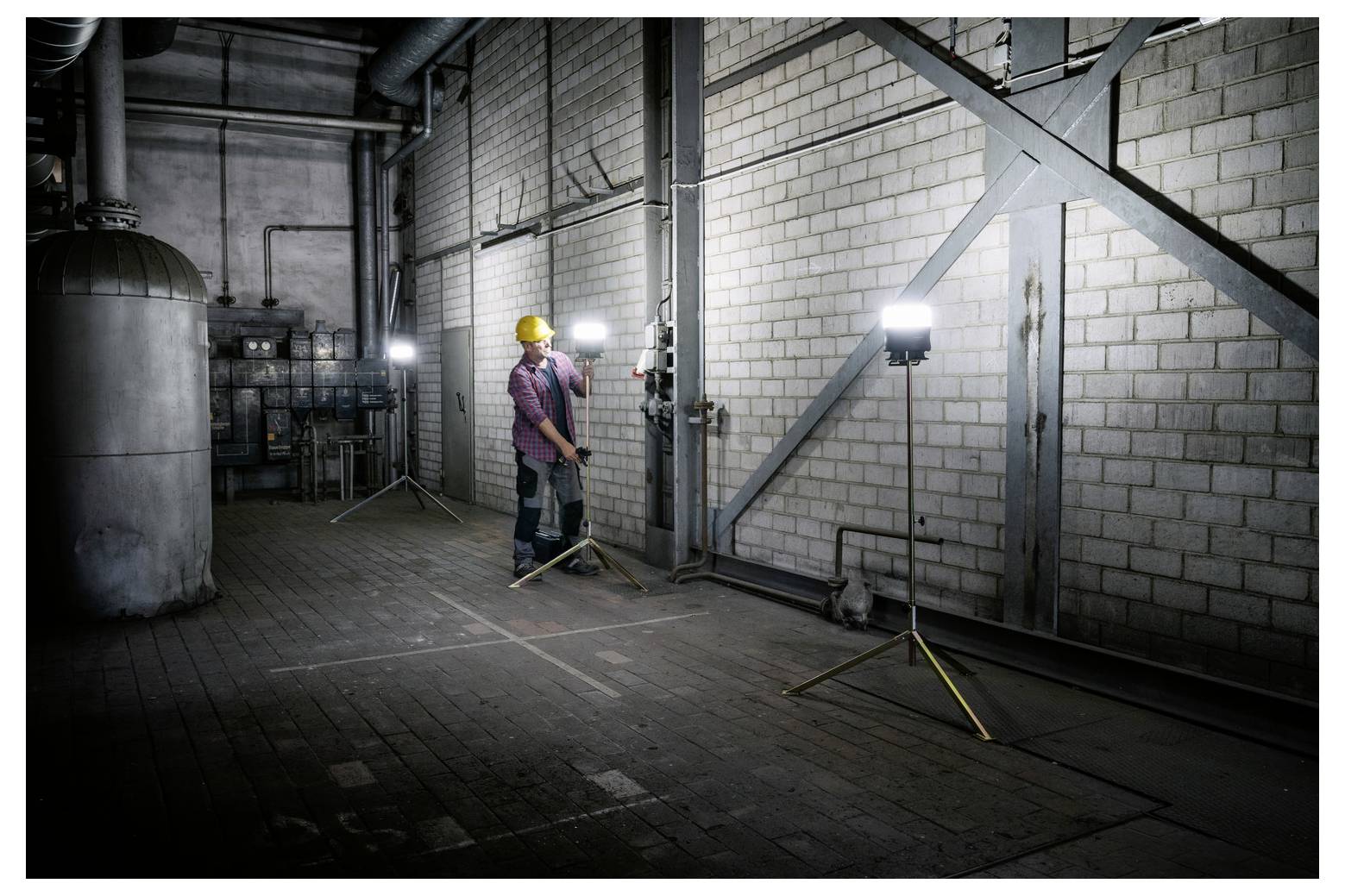 Arbeider in helm inspecteert buizen en kleppen in een donkere industriële kelder verlicht door draagbare werklichten.
