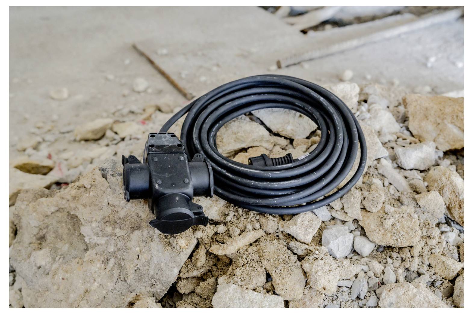 Opgerolde zwarte elektrische kabel met een stekker of connector die op gebroken beton en puin op een bouwplaats ligt.