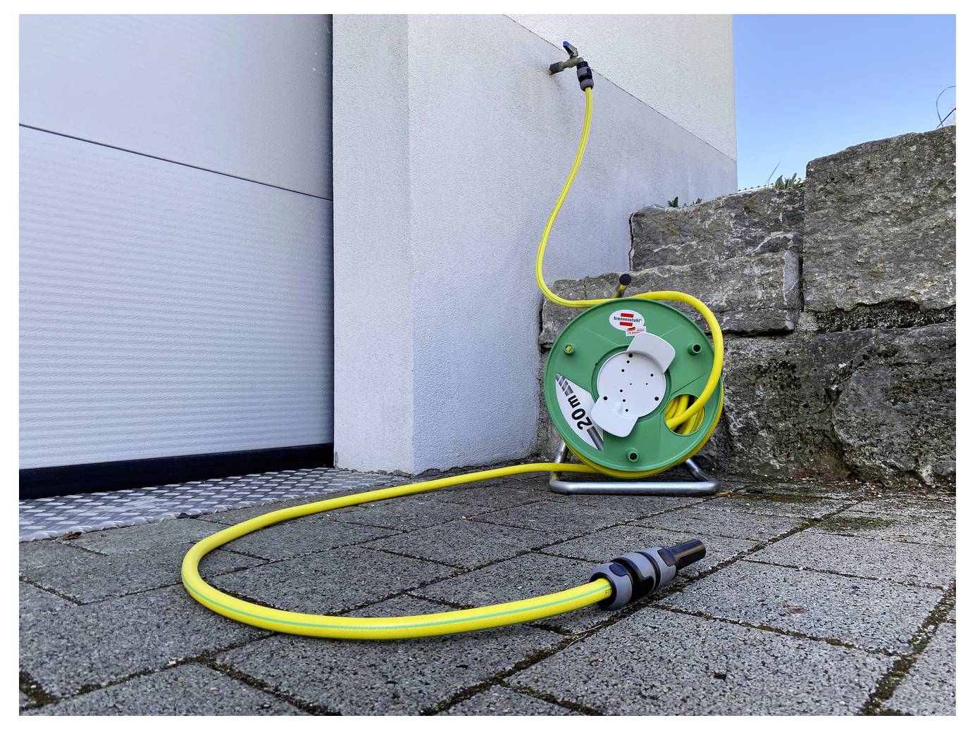 Gele tuinslang op een aan de muur gemonteerde haspel naast een garagedeur, met de slang uitgestrekt over een verharde patio dicht bij een stenen muur.