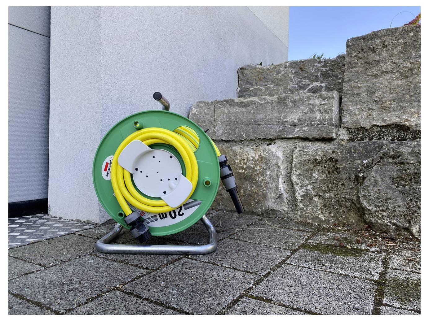 Groene slangenhaspel met opgerolde gele tuinslang en sproeikop, leunend naast een stenen muur op een geplaveide patio buiten.