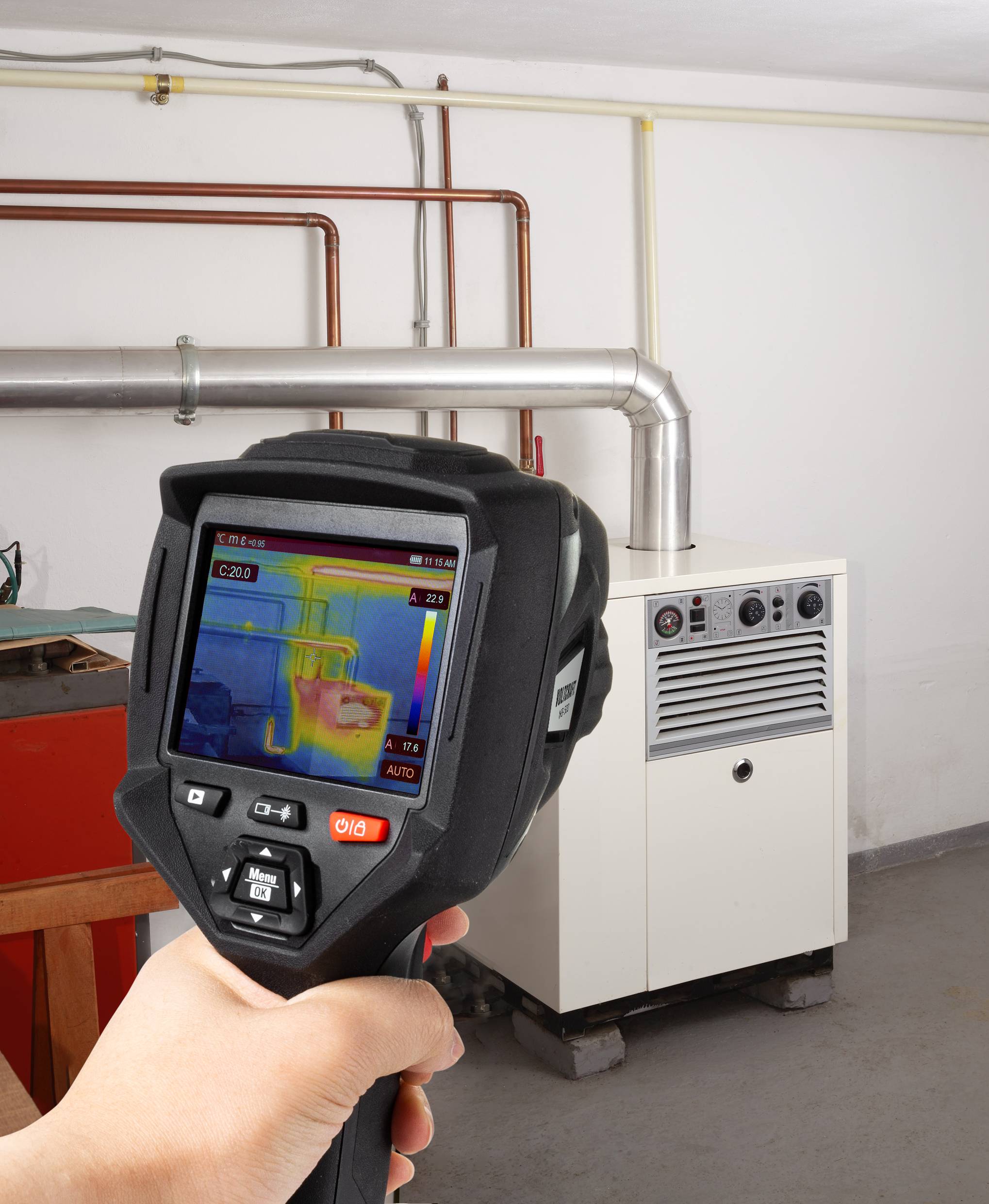 Een hand houdt een warmtebeeldcamera vast die verwarmingsleidingen in een kelderruimte met een warmtepomp inspecteert.